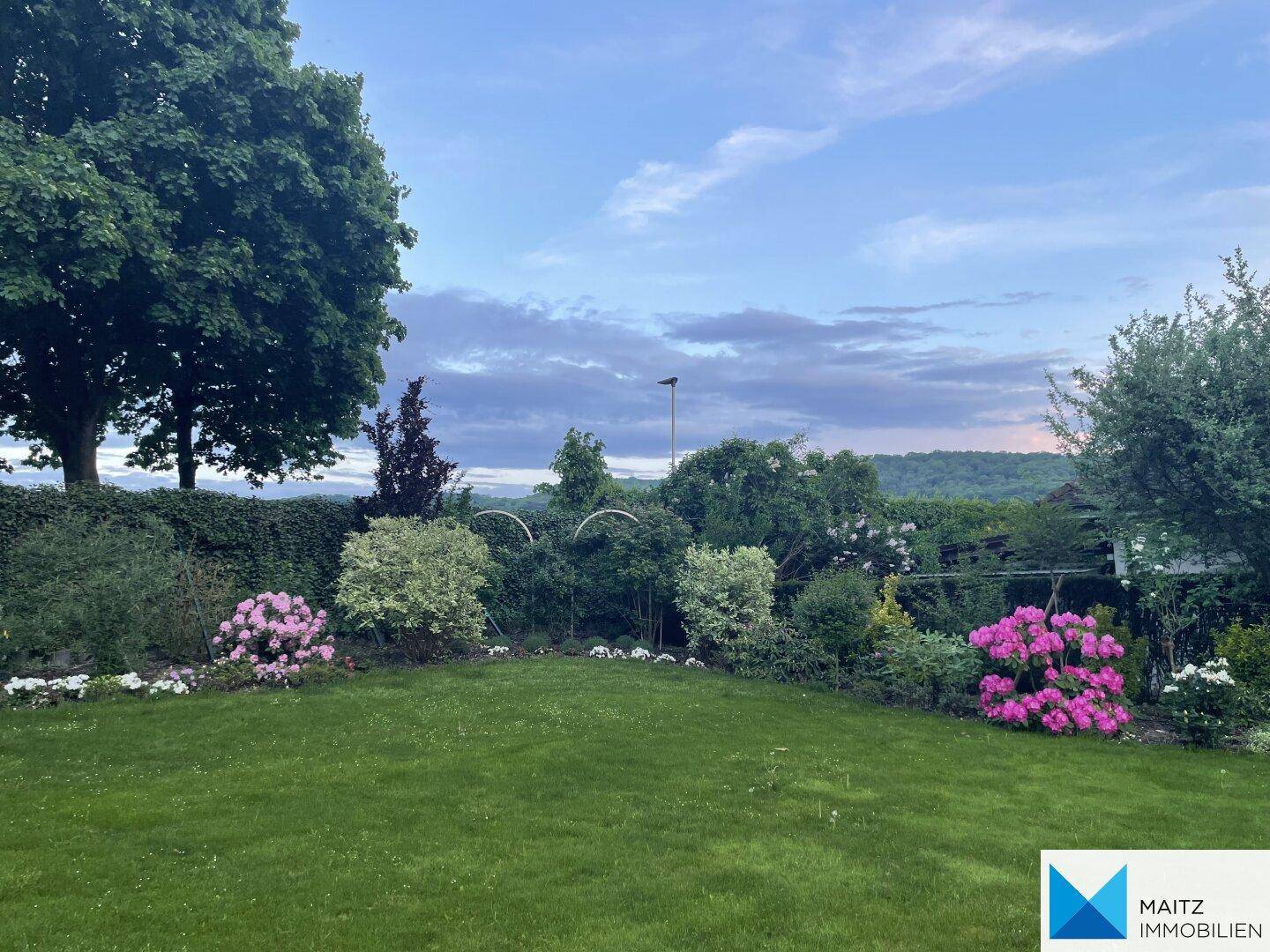 Weitläufiger Garten mit Rasenfläche und blühenden Sträuchern in der Abenddämmerung.