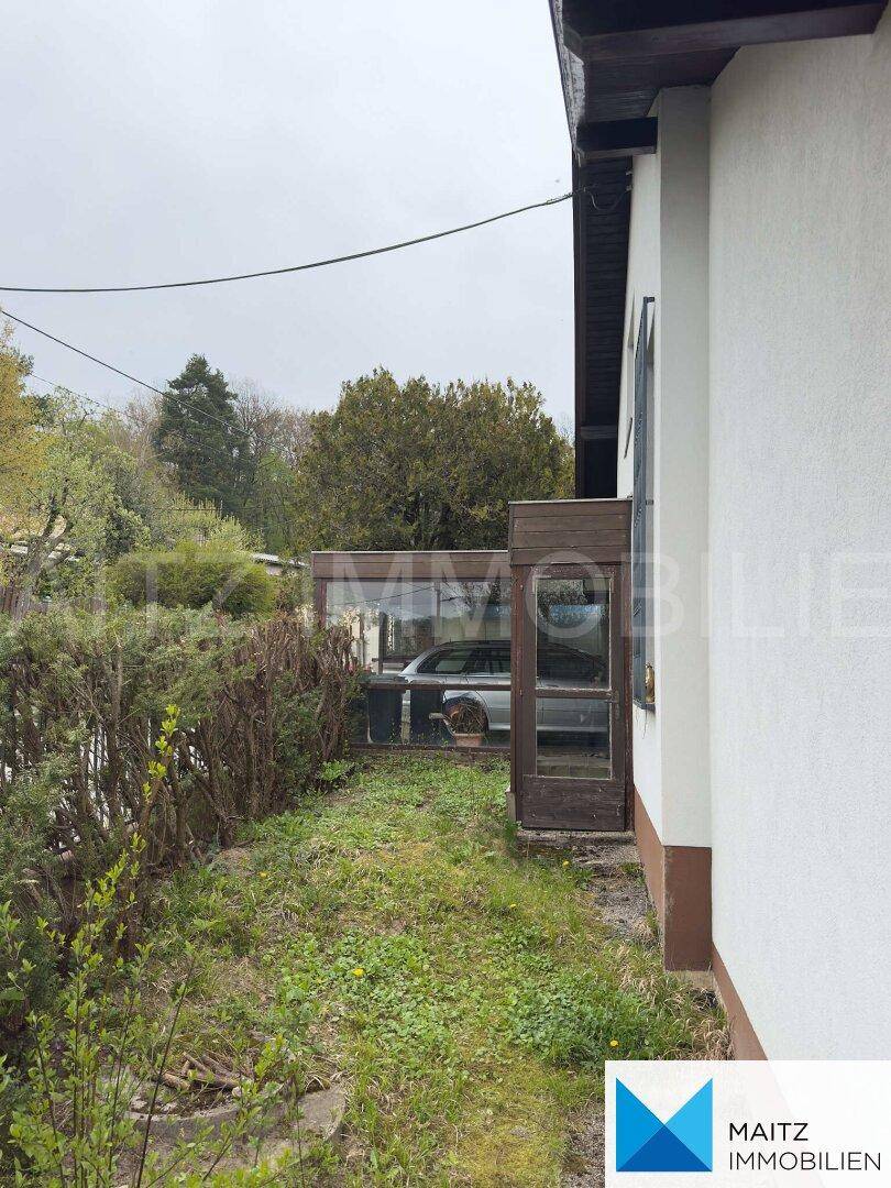 Seitenansicht des Hauses mit einer kleinen Glasstruktur und Hecke im Gartenbereich.