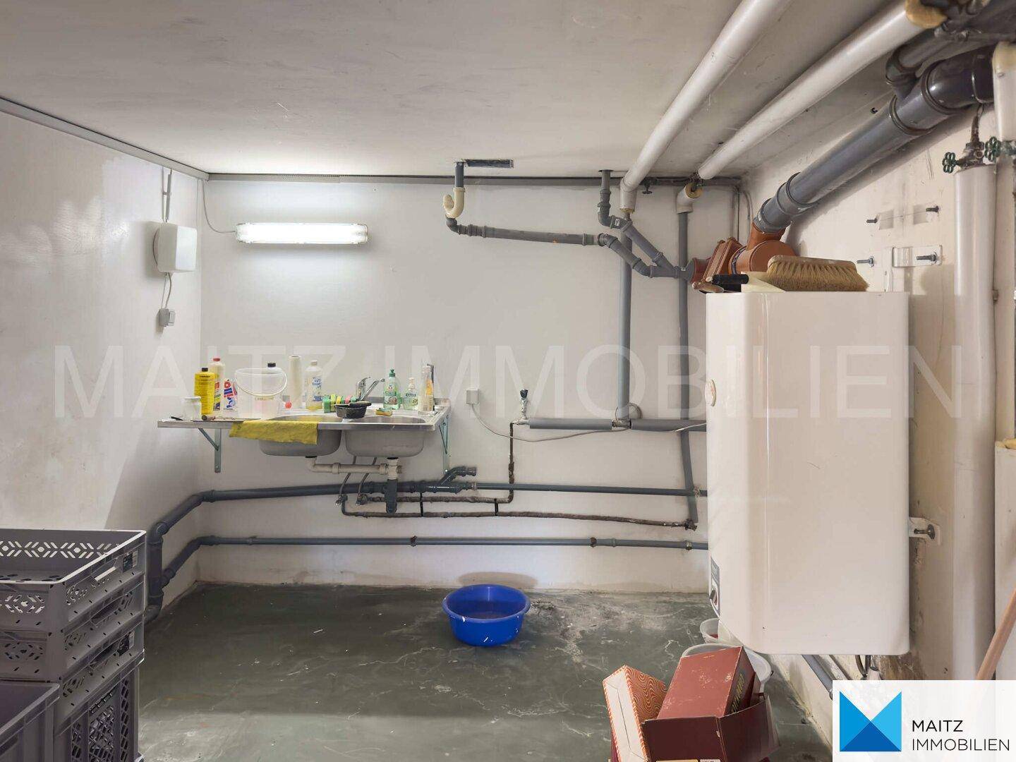 Funktionaler Hauswirtschaftsraum im Keller mit Waschbecken, Warmwasserbereiter und sichtbaren Leitungen.