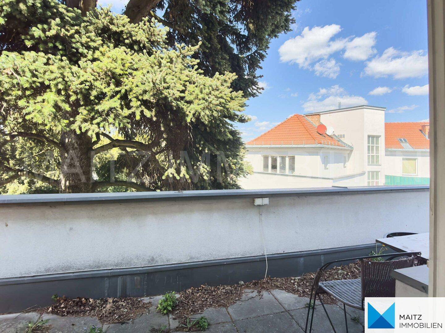 Außenbereich mit Blick auf grüne Bäume und die Dächer der Nachbargebäude, mit Platz für Sitzgelegenheiten.