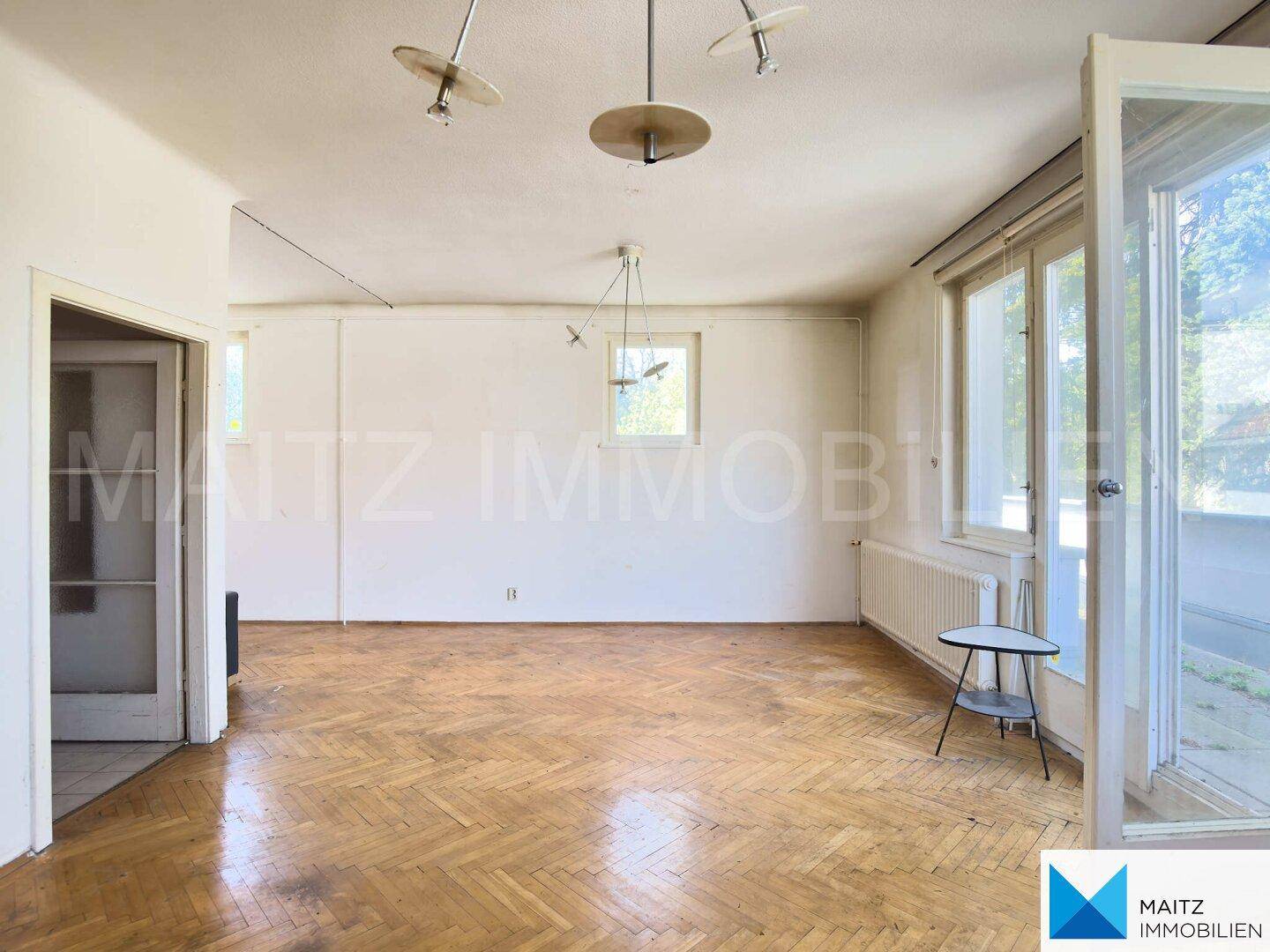 Geräumiges Wohnzimmer mit Fischgrätparkett, weißen Wänden und viel Tageslicht durch große Fenster.