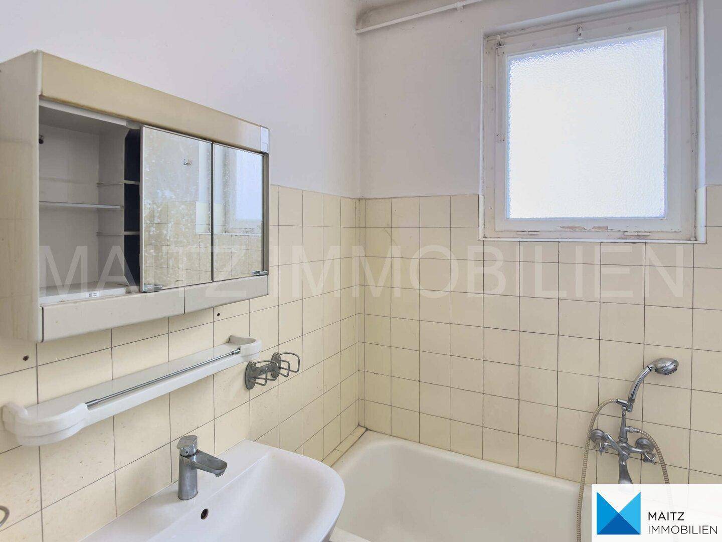 Badezimmer mit hellen Fliesen, Waschbecken, Spiegelschrank und Badewanne, mit Fenster für Tageslicht.