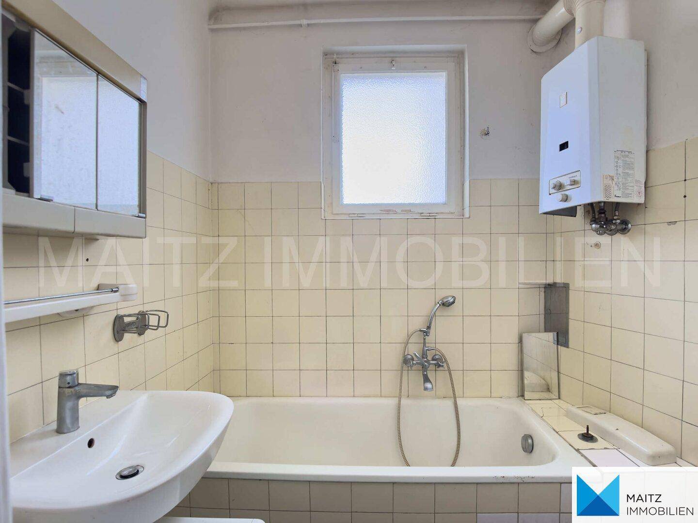 Badezimmer mit Badewanne, Waschbecken, Spiegelschrank und Gastherme, in einem älteren Zustand.