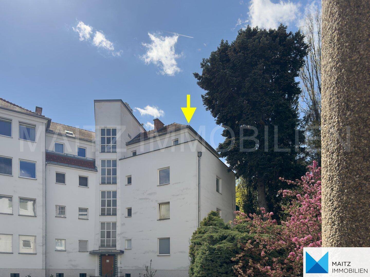Weißes Mehrfamilienhaus mit grüner Umgebung und einem Pfeil, der auf das Dach zeigt.