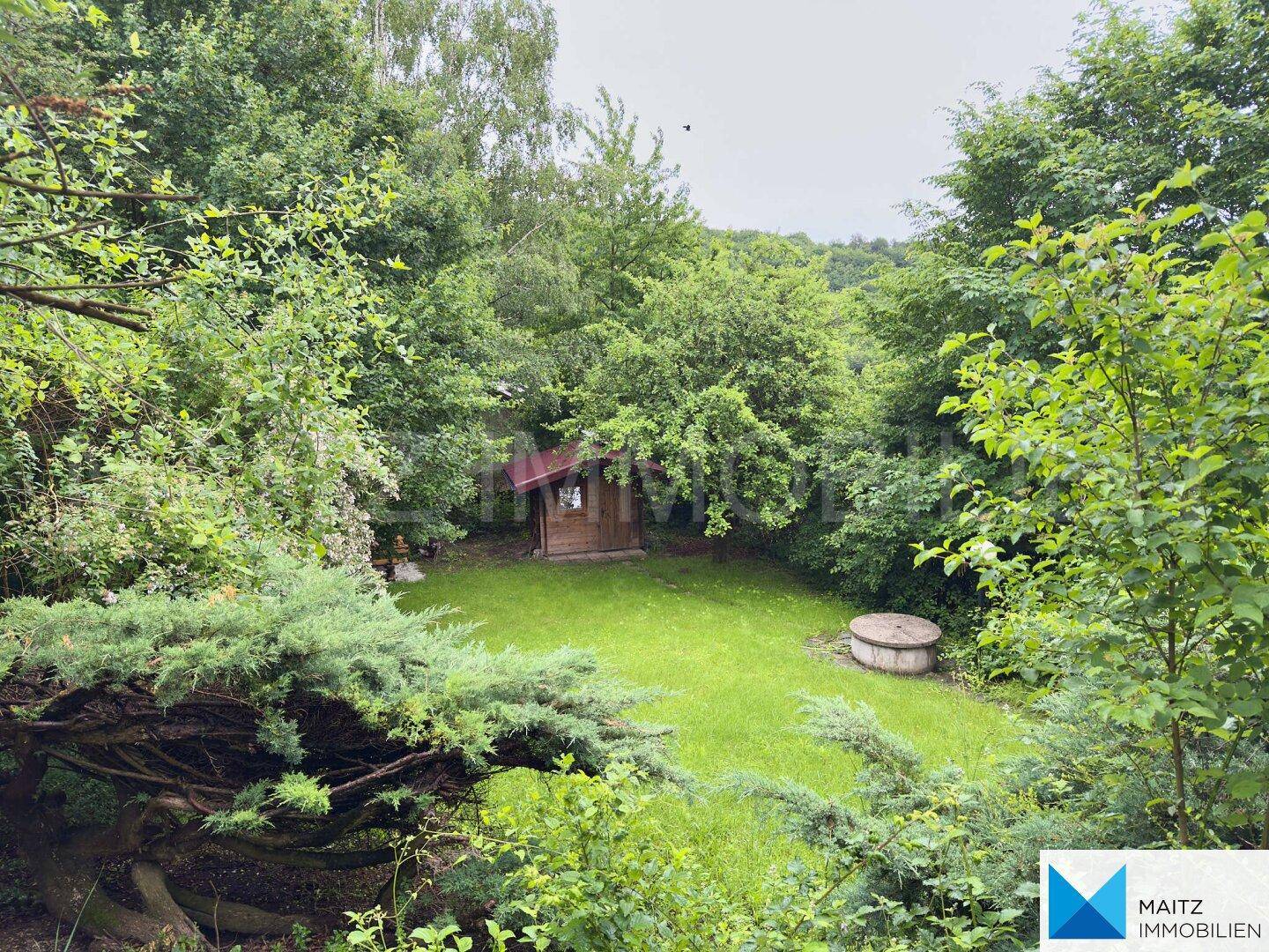 Idyllischer, grüner Garten mit Gartenhaus und altem Brunnen, umgeben von üppiger Vegetation.