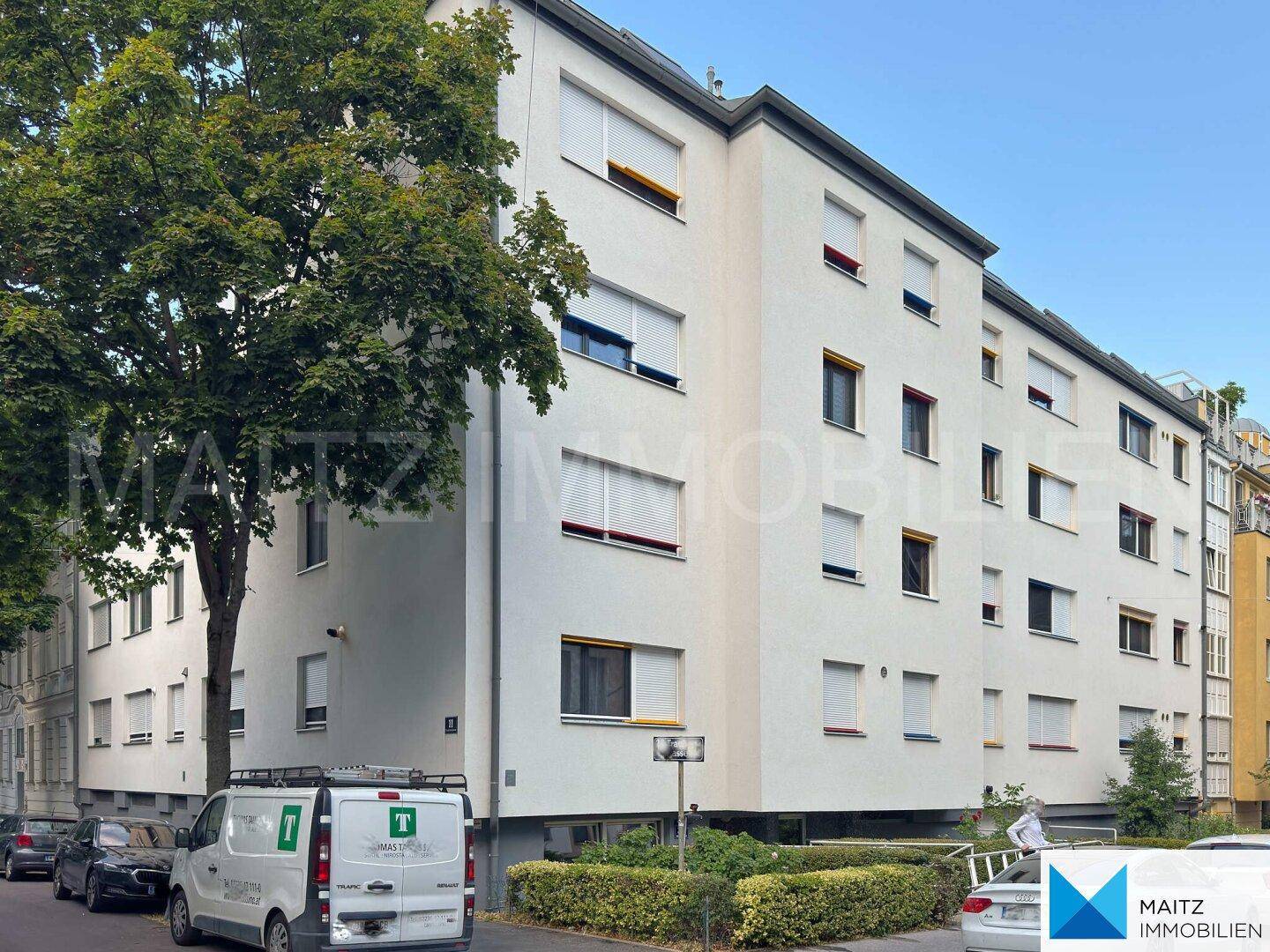 Mehrfamilienhaus mit weißer Fassade, vielen Fenstern und Bäumen an der Straße.