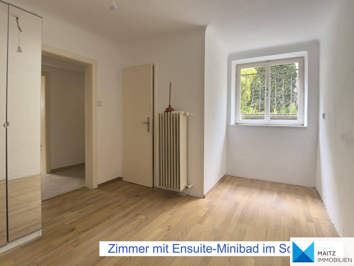 Helles Zimmer im Souterrain mit Parkettboden, Fenster und Zugang zu einem kleinen Bad.