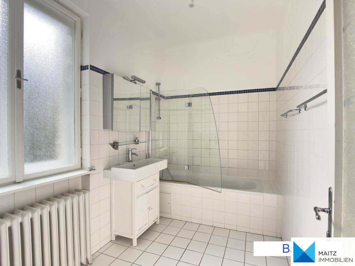 Badezimmer mit weißen Fliesen, Badewanne mit Duschwand und einem Waschbecken.