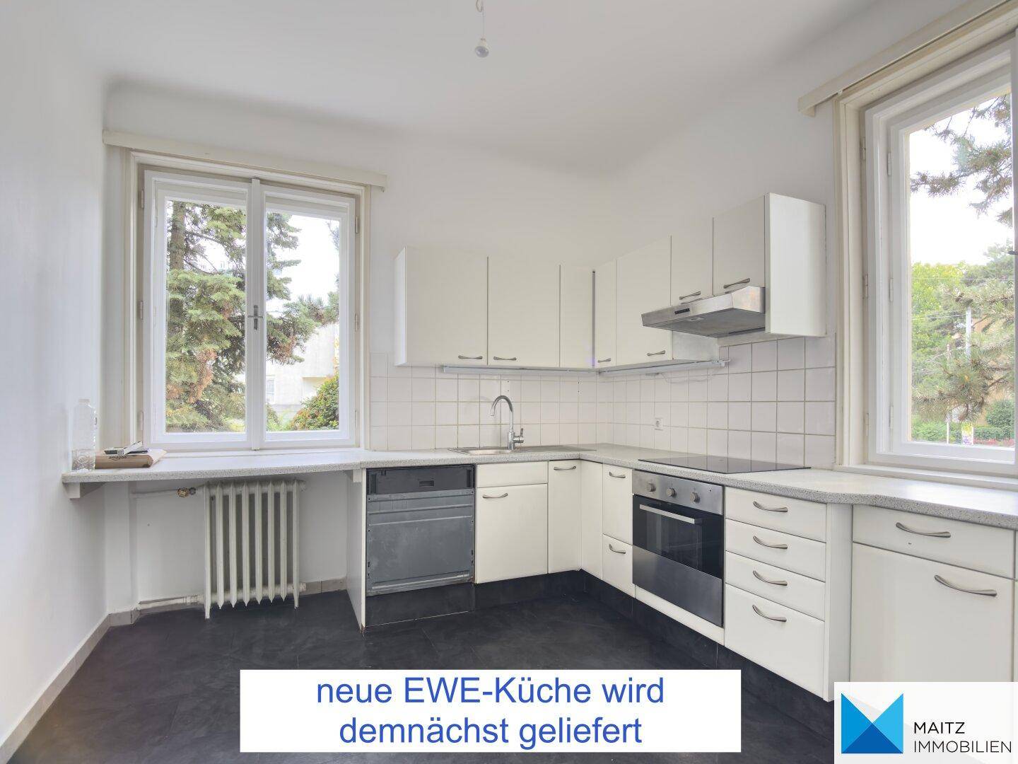 Helle Küche mit weißen Schränken, integrierten Geräten und Blick ins Grüne durch zwei Fenster.