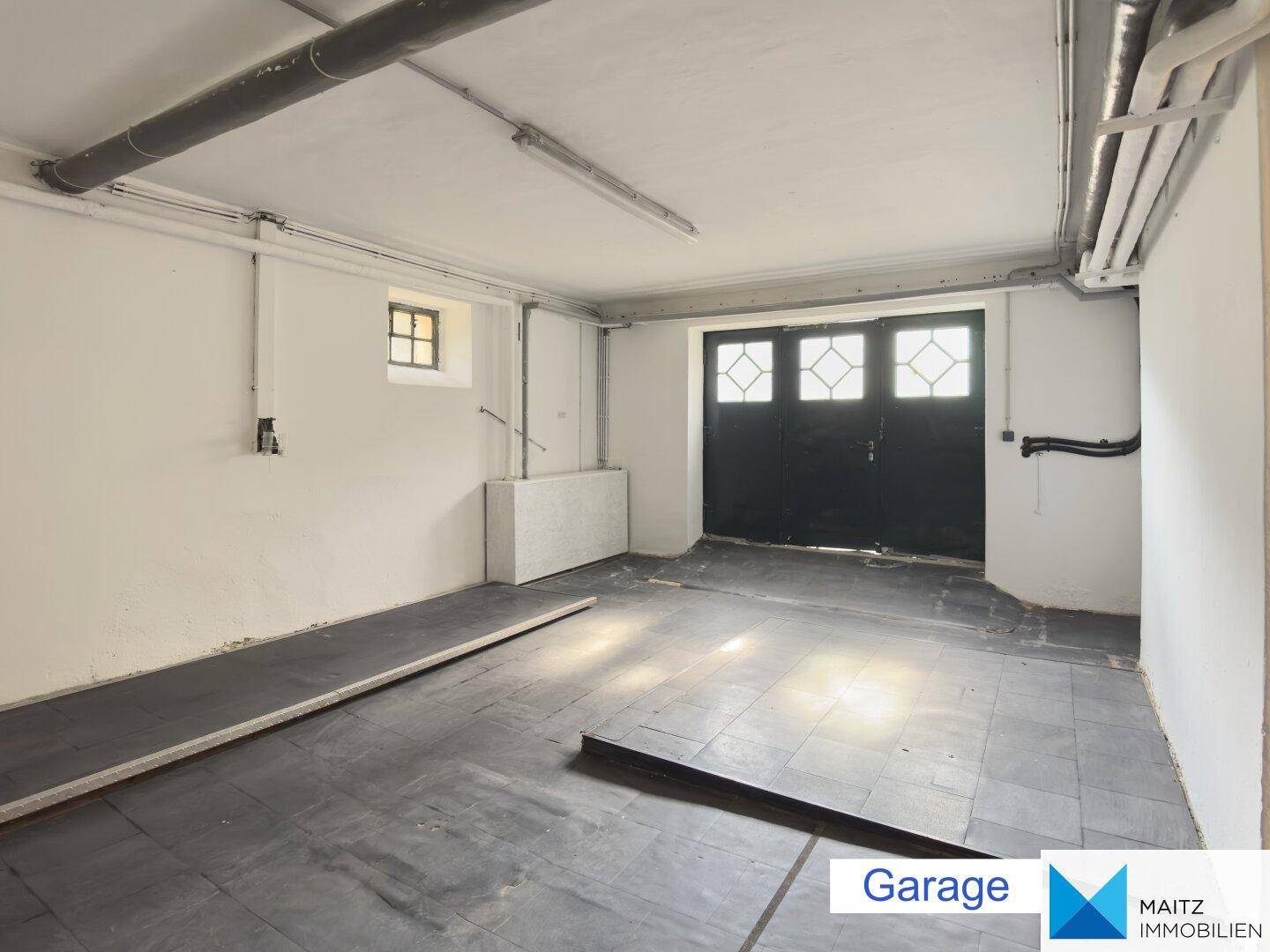 Geräumige Garage mit großen Doppeltoren, einem kleinen Fenster und sichtbaren Leitungen an der Decke.