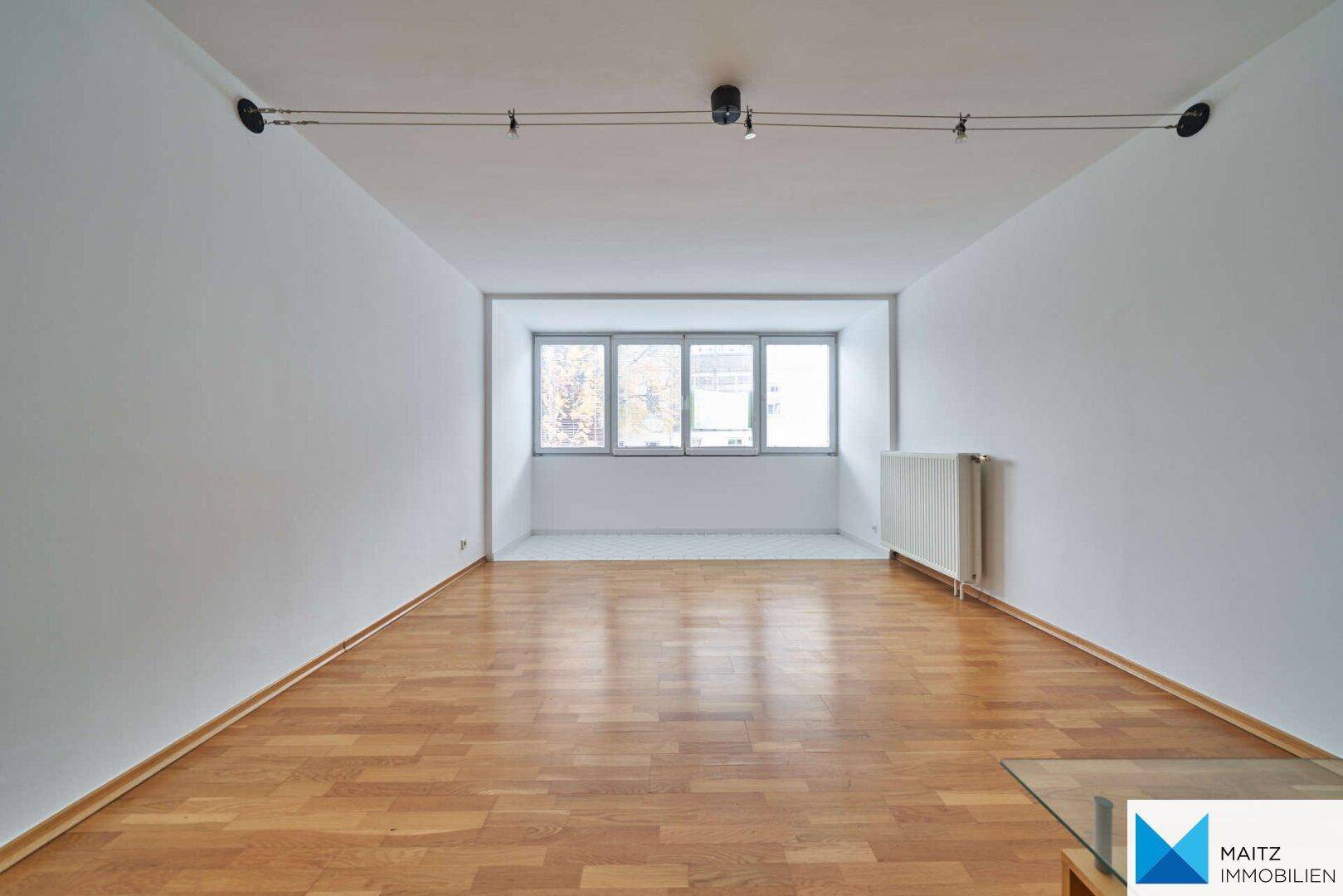 Heller, leerer Raum mit glänzendem Parkettboden und großen Fenstern, die viel Tageslicht hereinlassen.