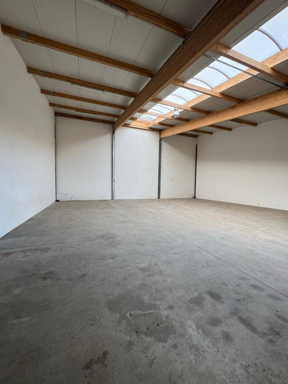 Geräumige, leere Halle mit Betonboden, hohen Decken und sichtbaren Holzträgern sowie Oberlichtern.