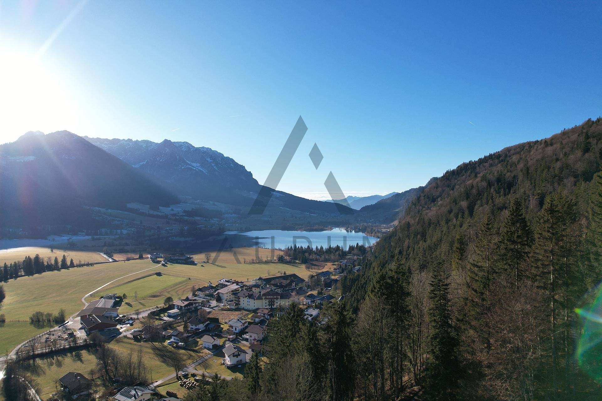Weitläufige Luftaufnahme eines malerischen Tals mit See, Bergen und einem kleinen Dorf unter strahlend blauem Himmel.