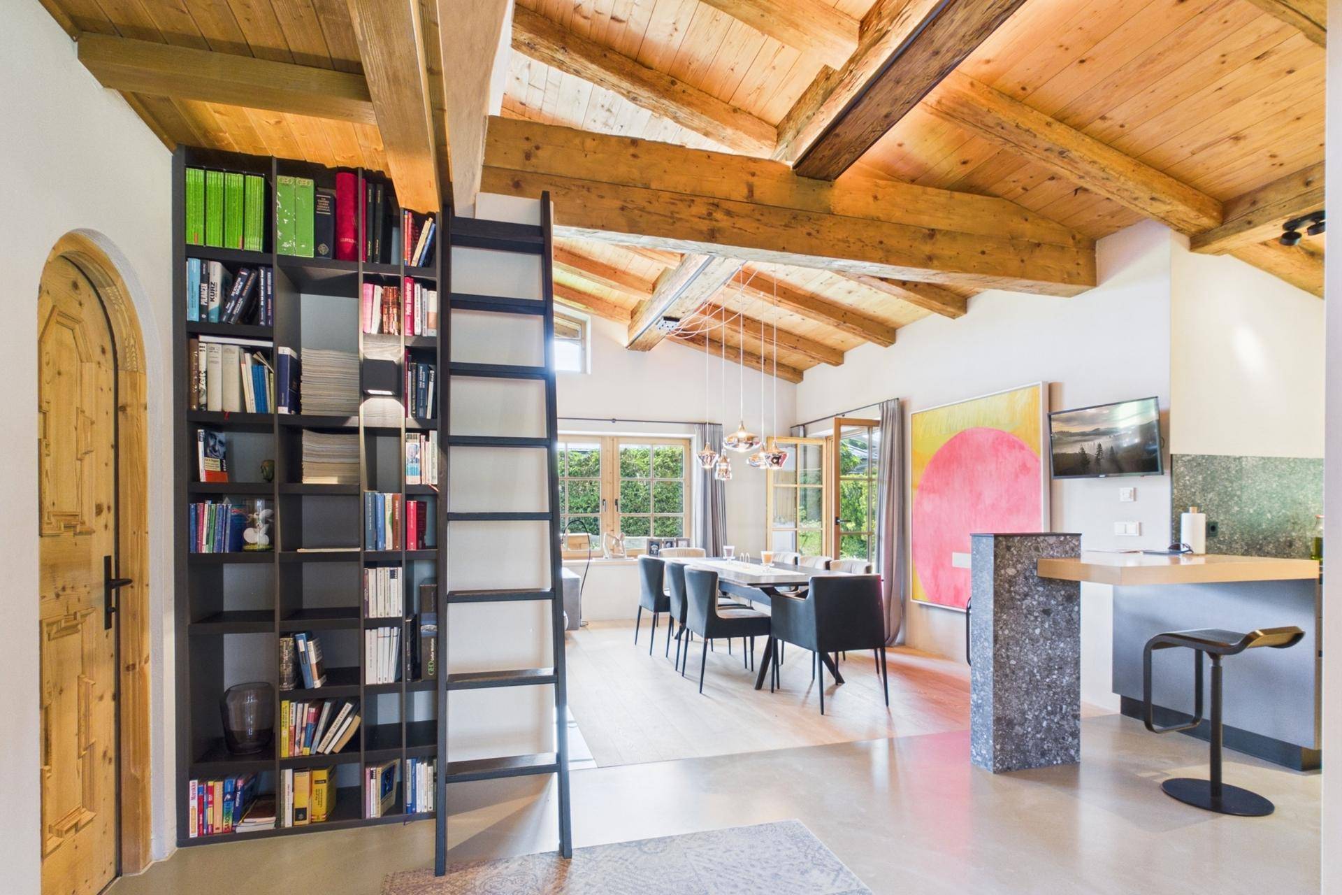 Heller Wohn- und Essbereich mit hoher Decke, Holzbalken und integriertem Bücherregal.