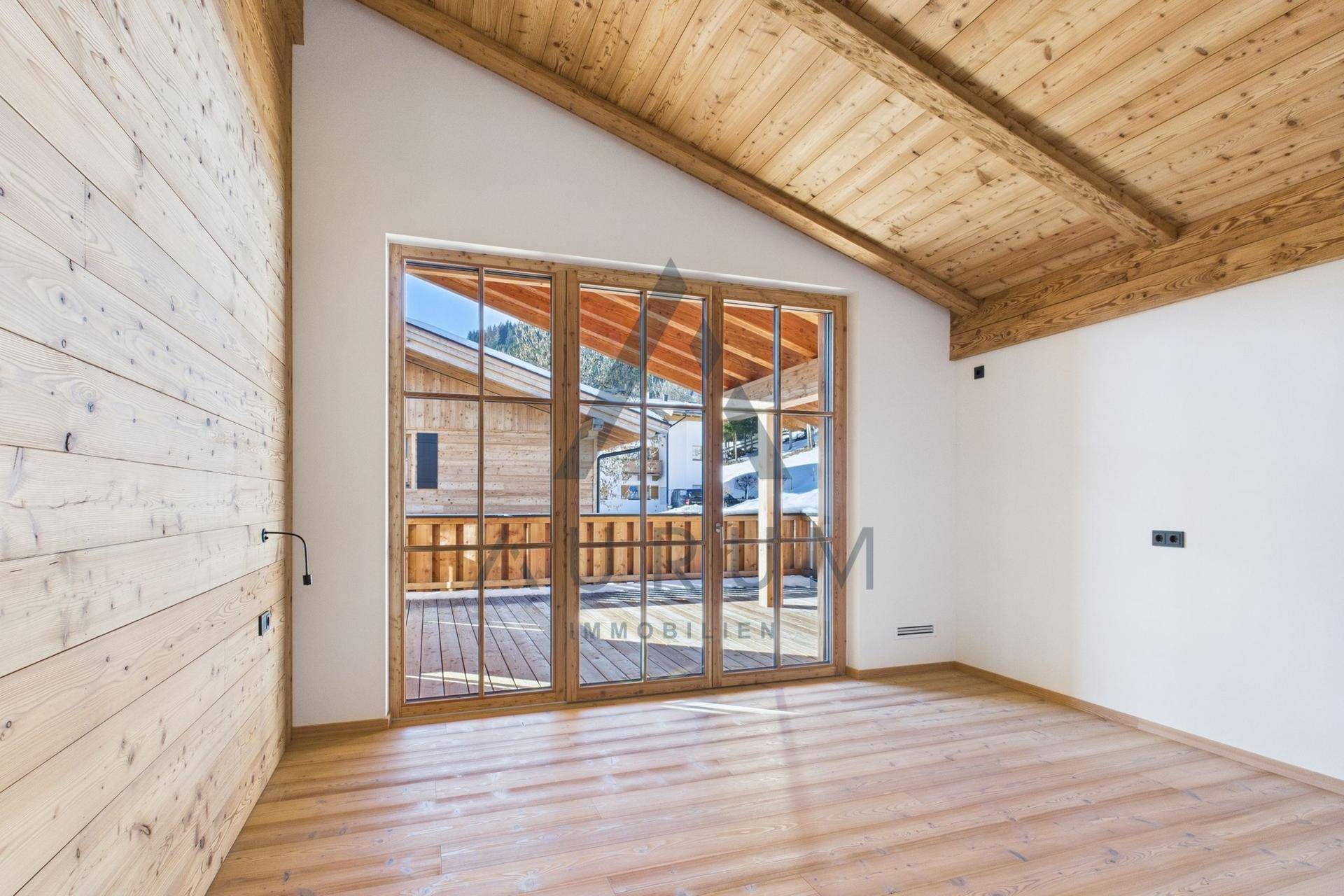 Heller, leerer Raum mit Holzwänden, Dachschräge und großen Fenstern zum Balkon.