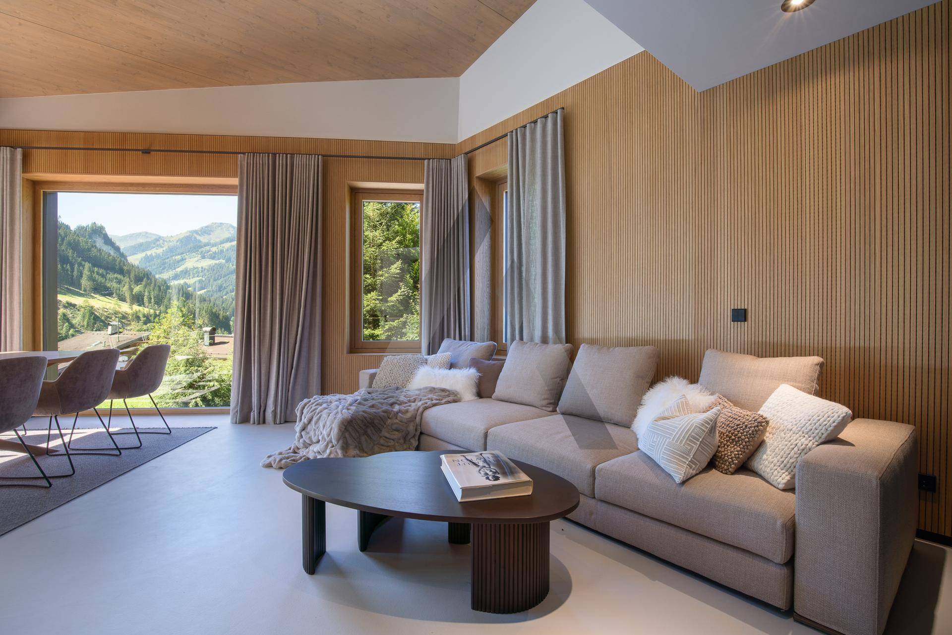 Lichtdurchflutetes Wohnzimmer mit großem Sofa, Holzwänden und weitem Bergblick durch Panoramafenster.