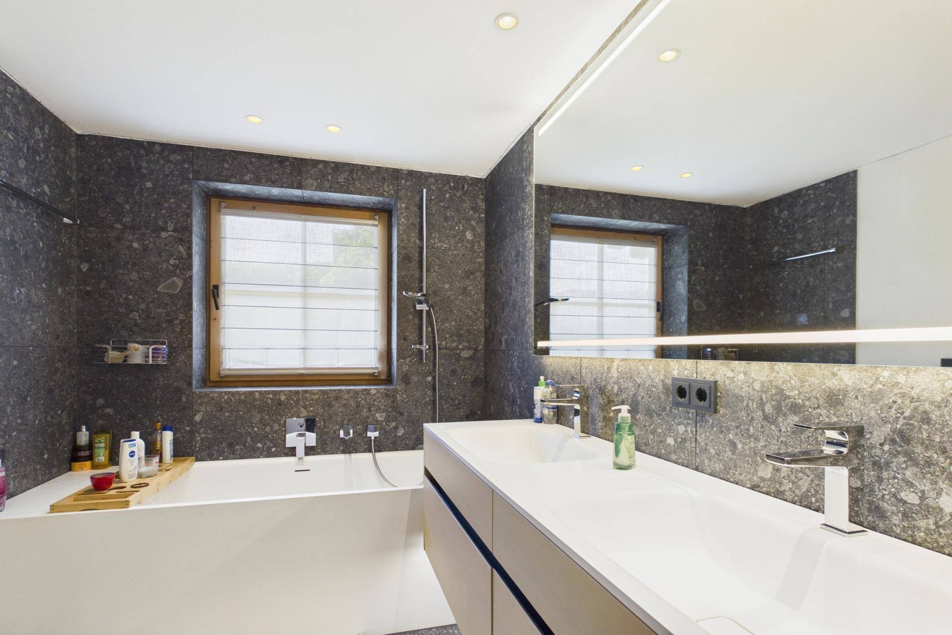Modernes Badezimmer mit dunklen Fliesen, Badewanne, Doppelwaschbecken und Fenster.