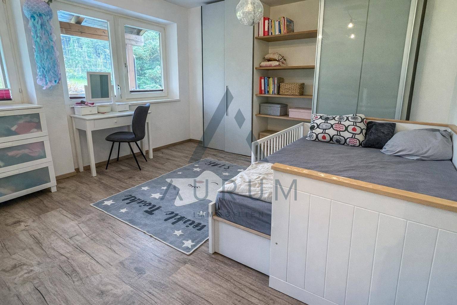 Kinderzimmer mit hellem Holzboden, Schreibtisch und großem Fenster mit Blick ins Grüne.