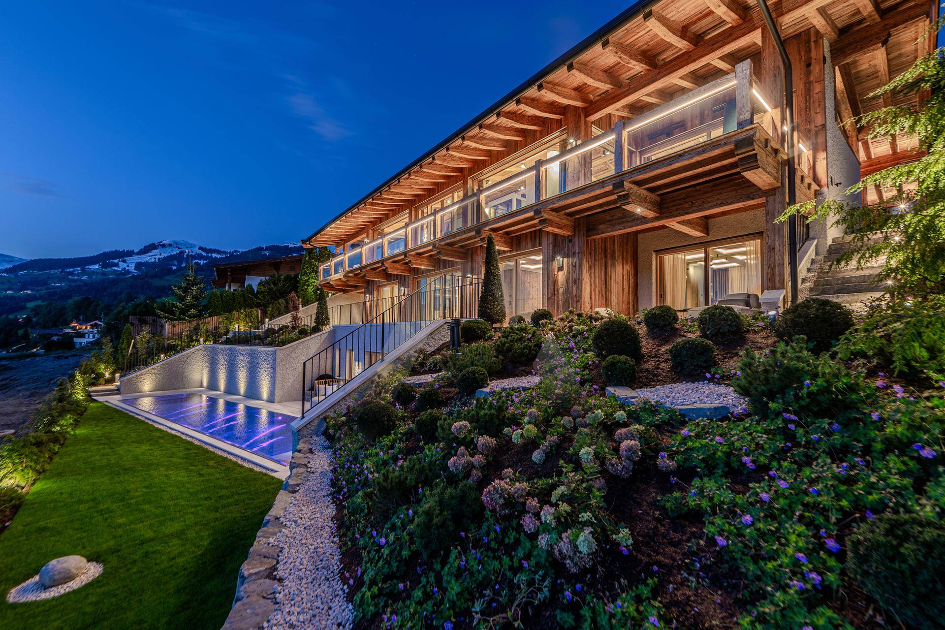 Beeindruckende moderne Chalet-Architektur mit beleuchteten Terrassen, Pool und gepflegtem Gartenbereich am Abend.