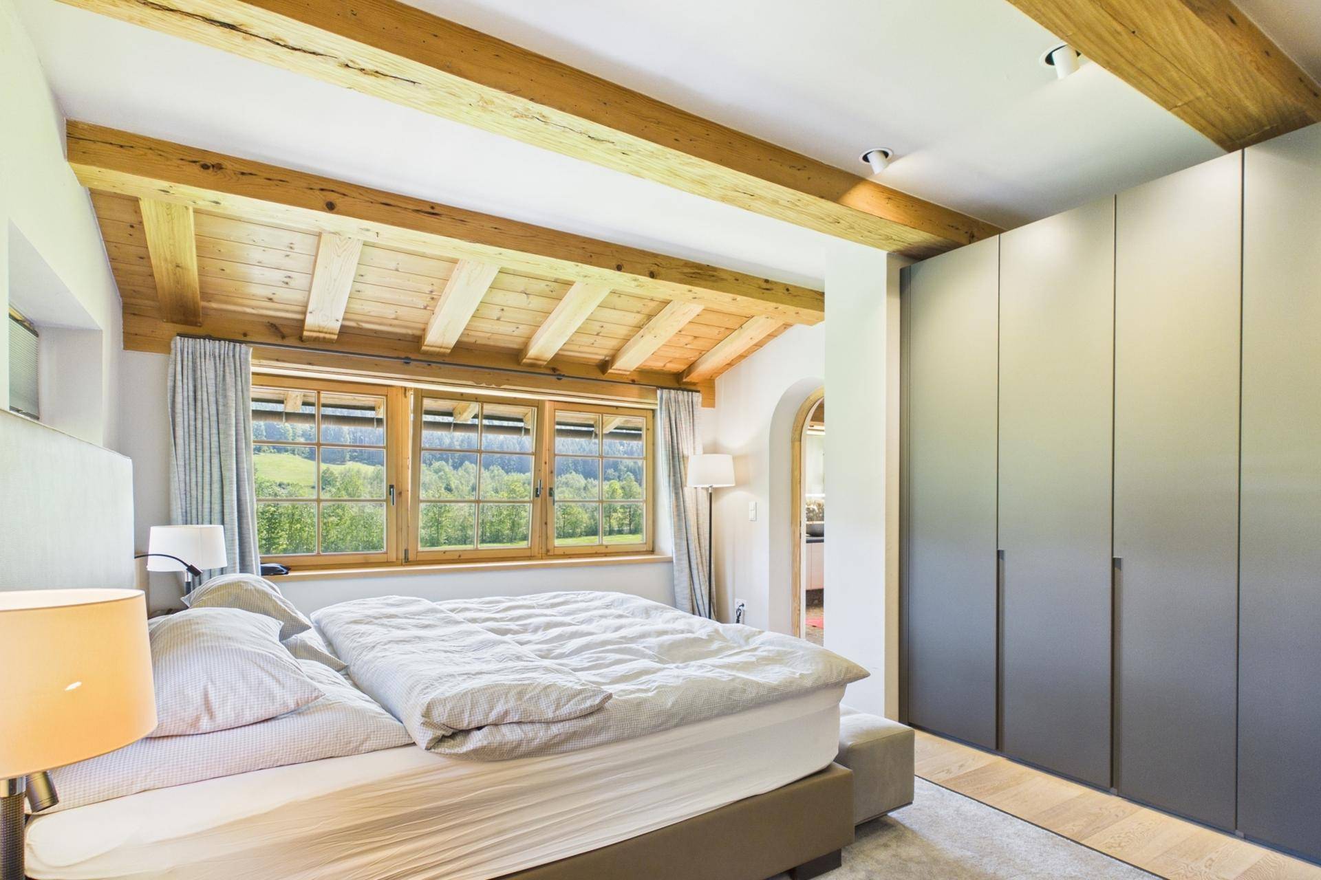 Helles Schlafzimmer mit Holzbalkendecke, Einbauschränken und Blick in die Natur.