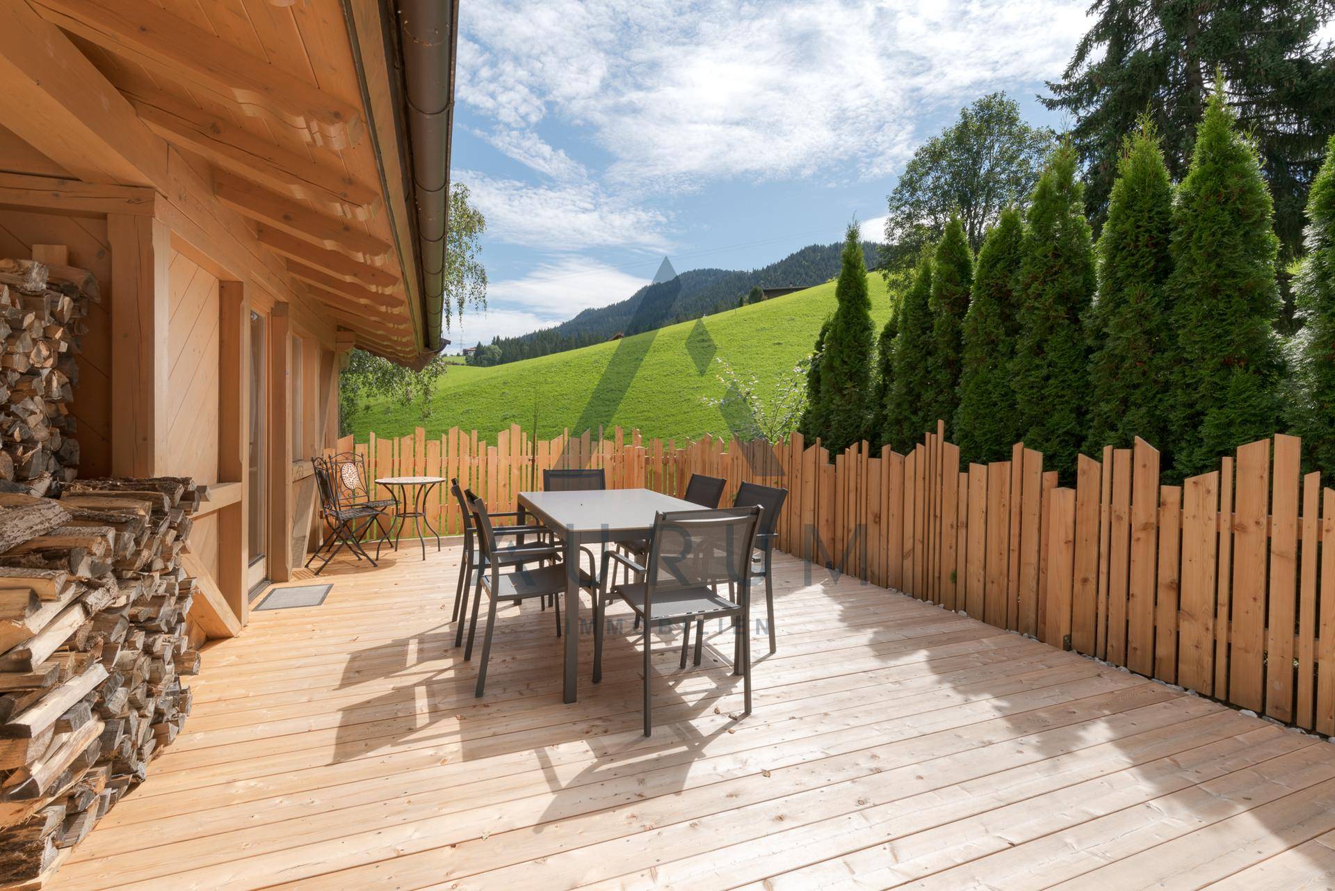 Geräumige Holzterrasse mit Essbereich im Freien, umgeben von grünen Hügeln und Bäumen.