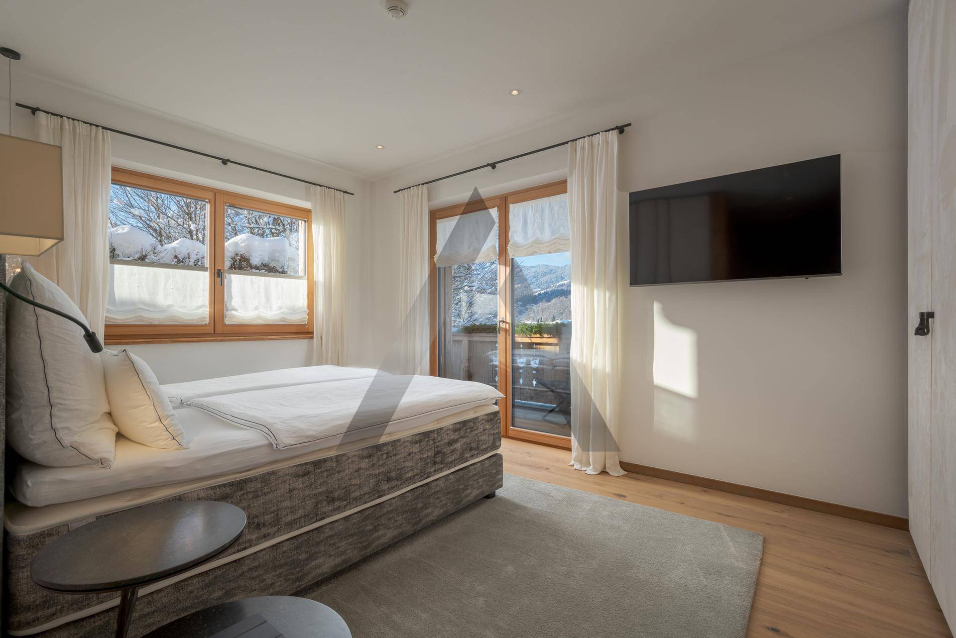 Helles Schlafzimmer mit Doppelbett, Holzboden und Zugang zu einem sonnigen Balkon mit Bergblick.