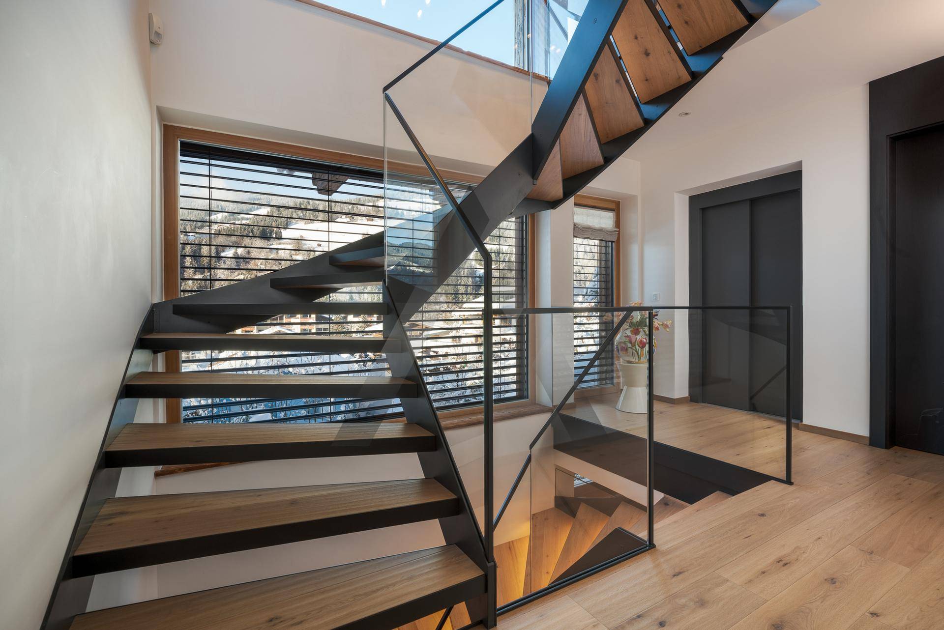 Elegante Holztreppe mit Glasgeländer und Blick auf die verschneite Berglandschaft, lichtdurchflutet.