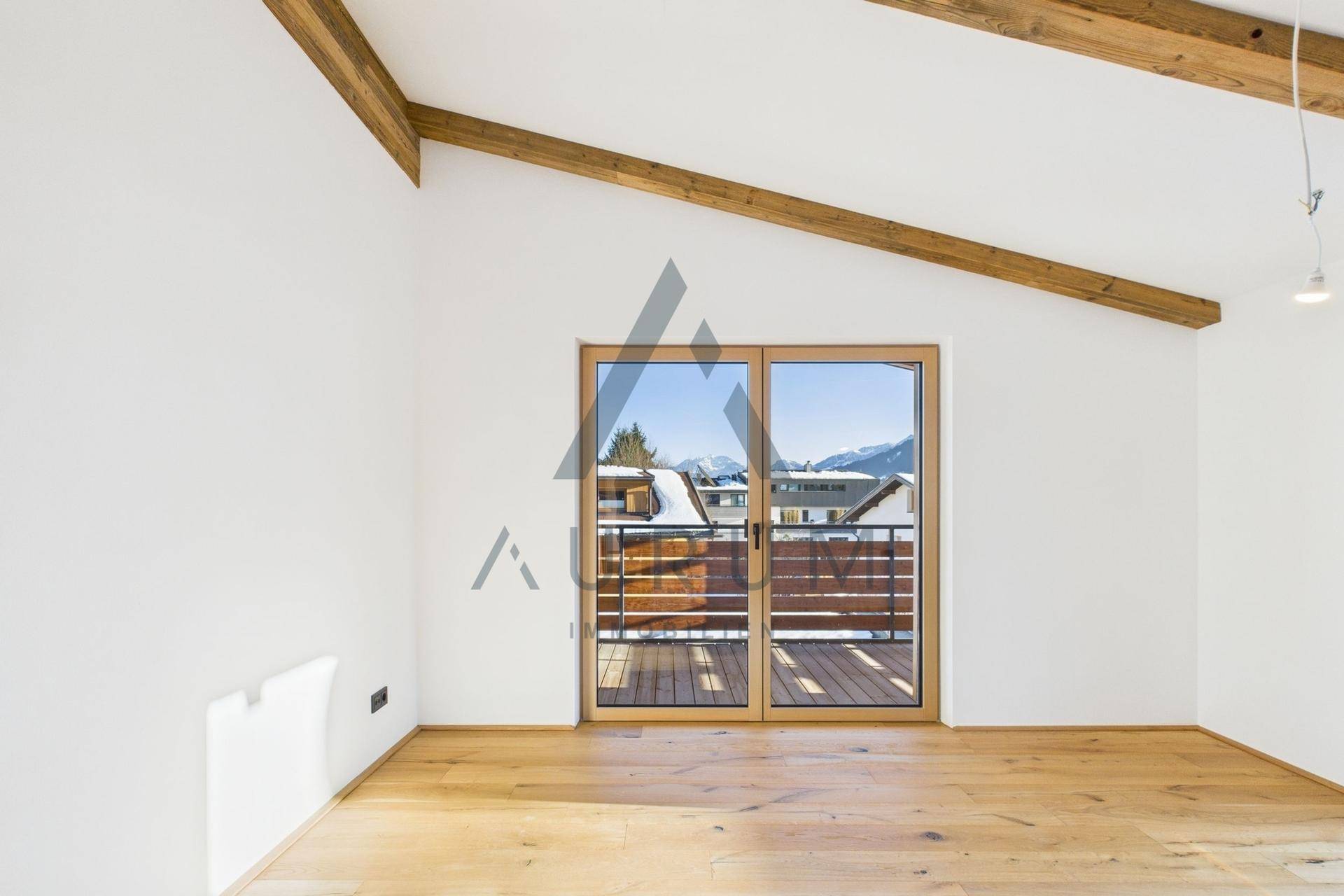 Geräumiger, heller Raum mit Holzboden, sichtbaren Balken und Zugang zu einem sonnigen Balkon.