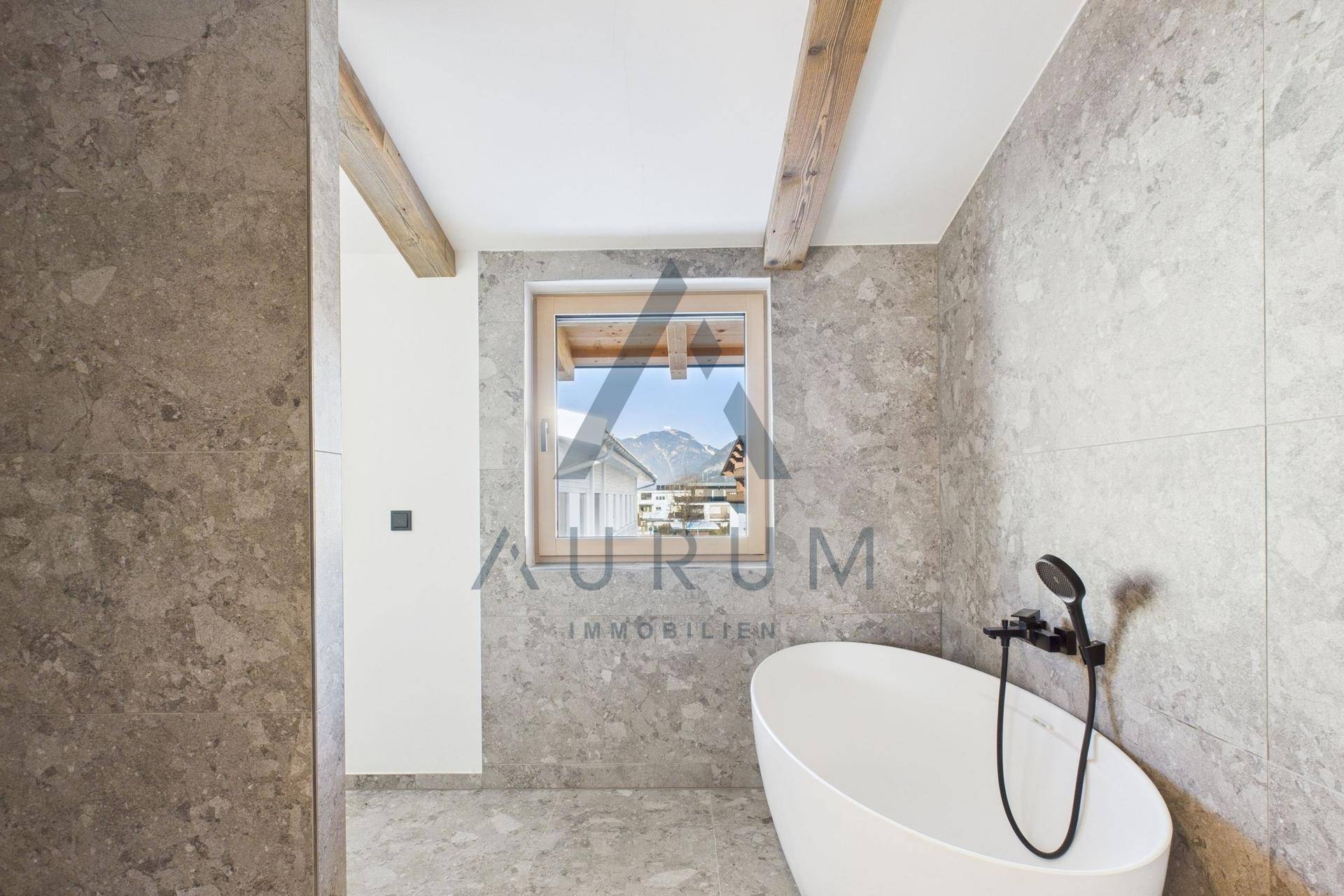 Elegantes Badezimmer mit freistehender Badewanne, grauen Fliesen und einem Fenster mit Bergblick.