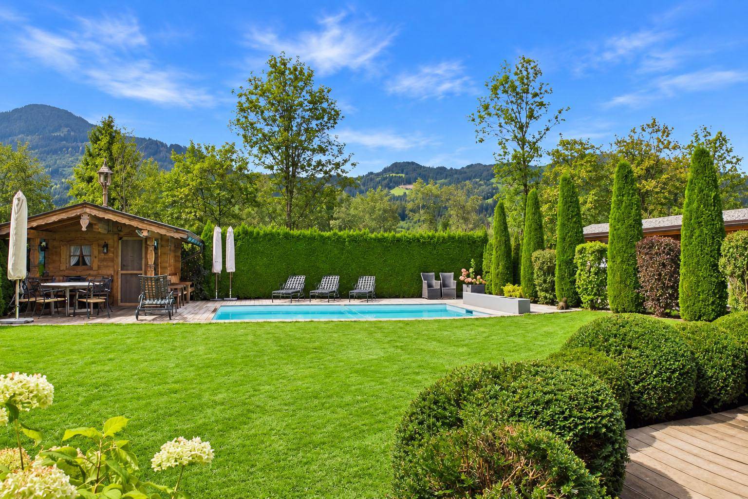 Weitläufiger Garten mit Pool, Liegestühlen und einer gemütlichen Gartenhütte vor Bergkulisse.