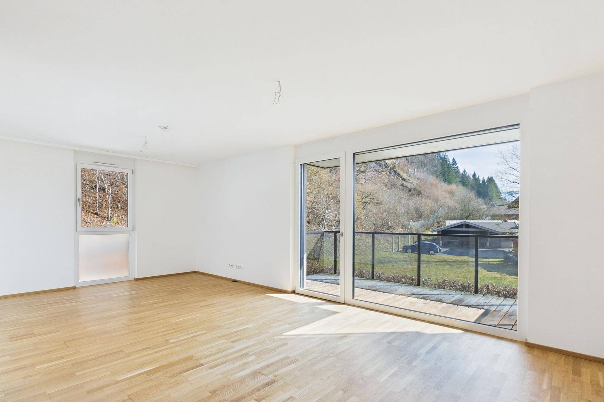 Geräumiges, leeres Wohnzimmer mit großen Fenstern, Holzboden und Blick auf die umliegende Natur.