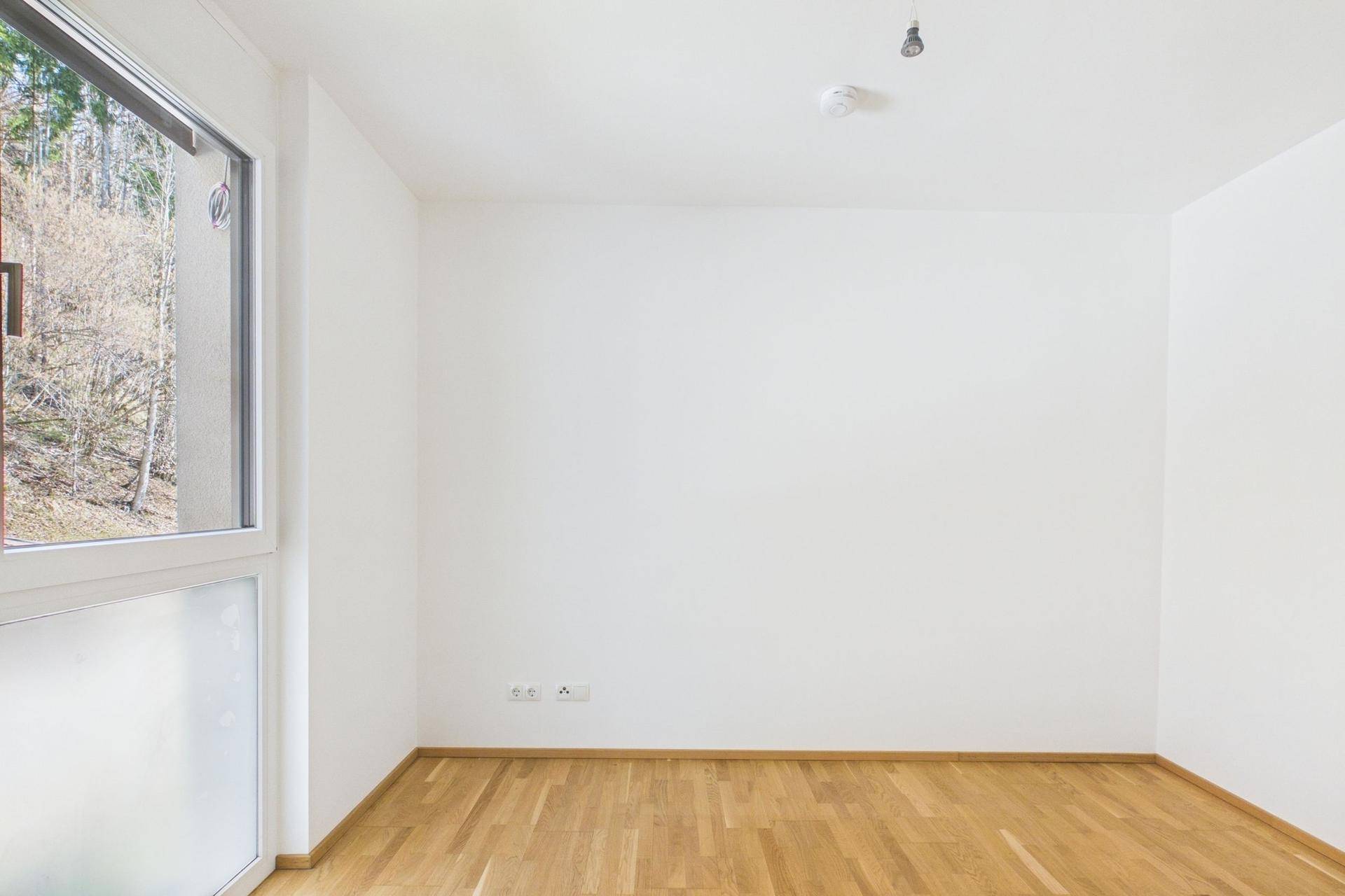 Leeres Schlafzimmer mit Holzboden und einem Fenster, das viel Tageslicht hereinlässt.
