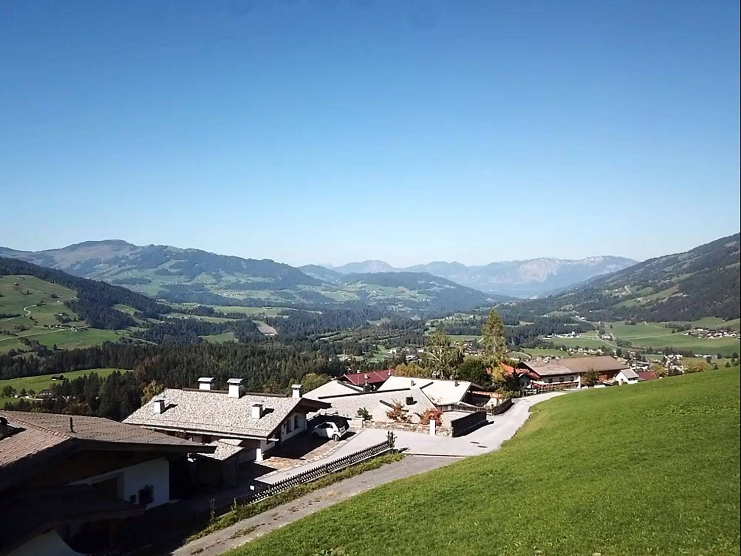 Blick über ein Tal mit Häusern, grünen Hügeln und einer Bergkette am Horizont unter klarem Himmel.