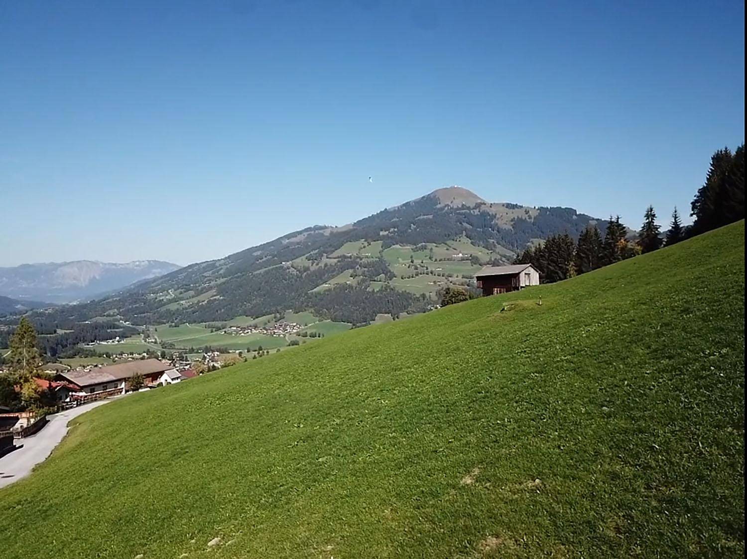 Grüne Hügellandschaft mit vereinzelten Häusern und Bergen im Hintergrund unter blauem Himmel.