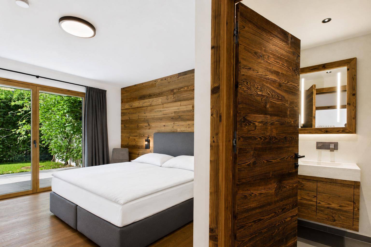 Schlafzimmer mit Holzwand, Doppelbett und Blick auf den Garten, angrenzend an ein modernes Bad.