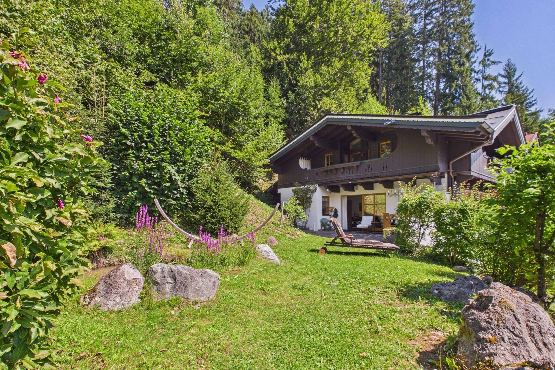 Gartenbereich mit Hängematte und Liegestuhl, umgeben von üppigem Grün und Blumen.