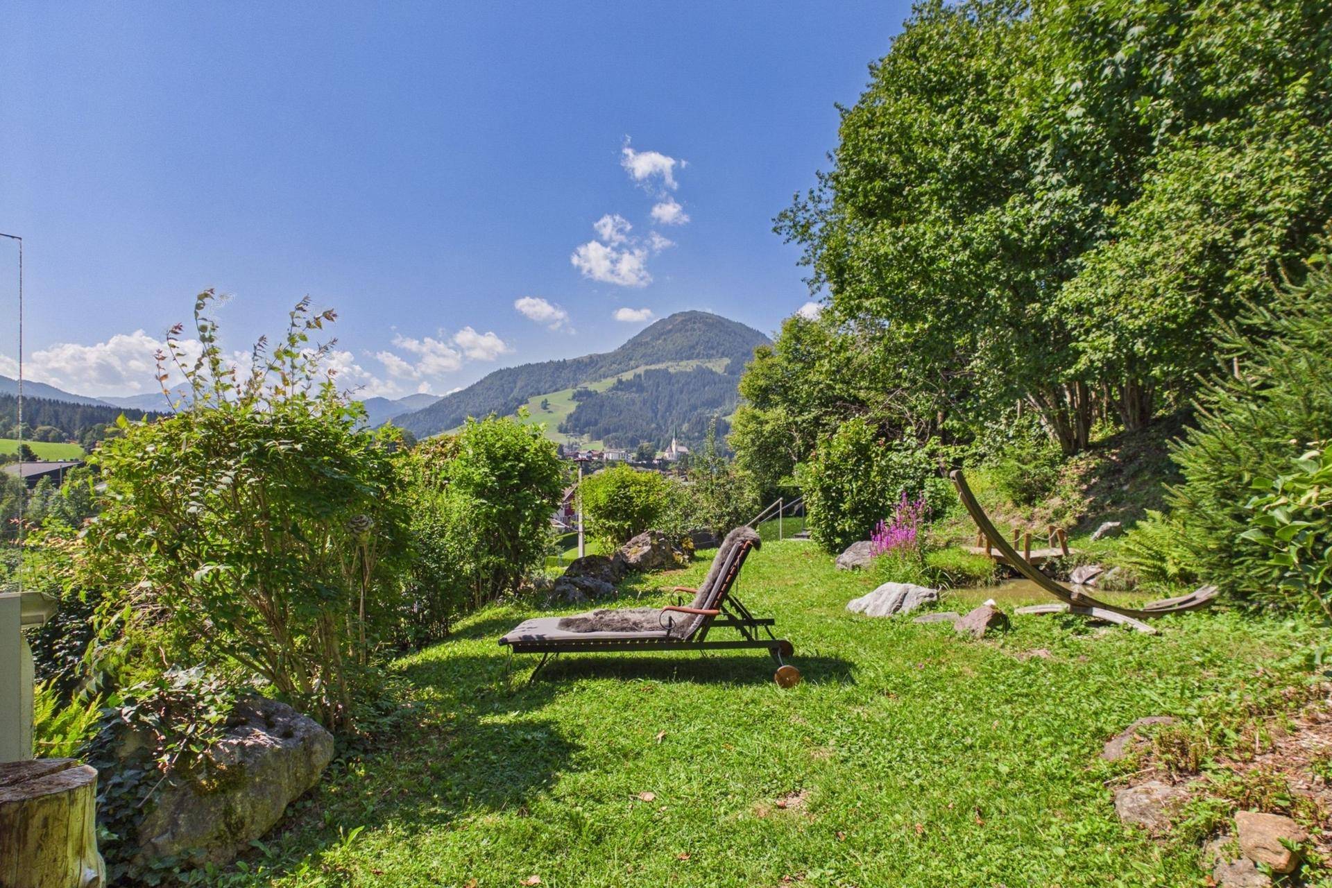 Gepflegter Gartenbereich mit Liegestuhl, umgeben von Bäumen und Sträuchern, mit Bergblick.