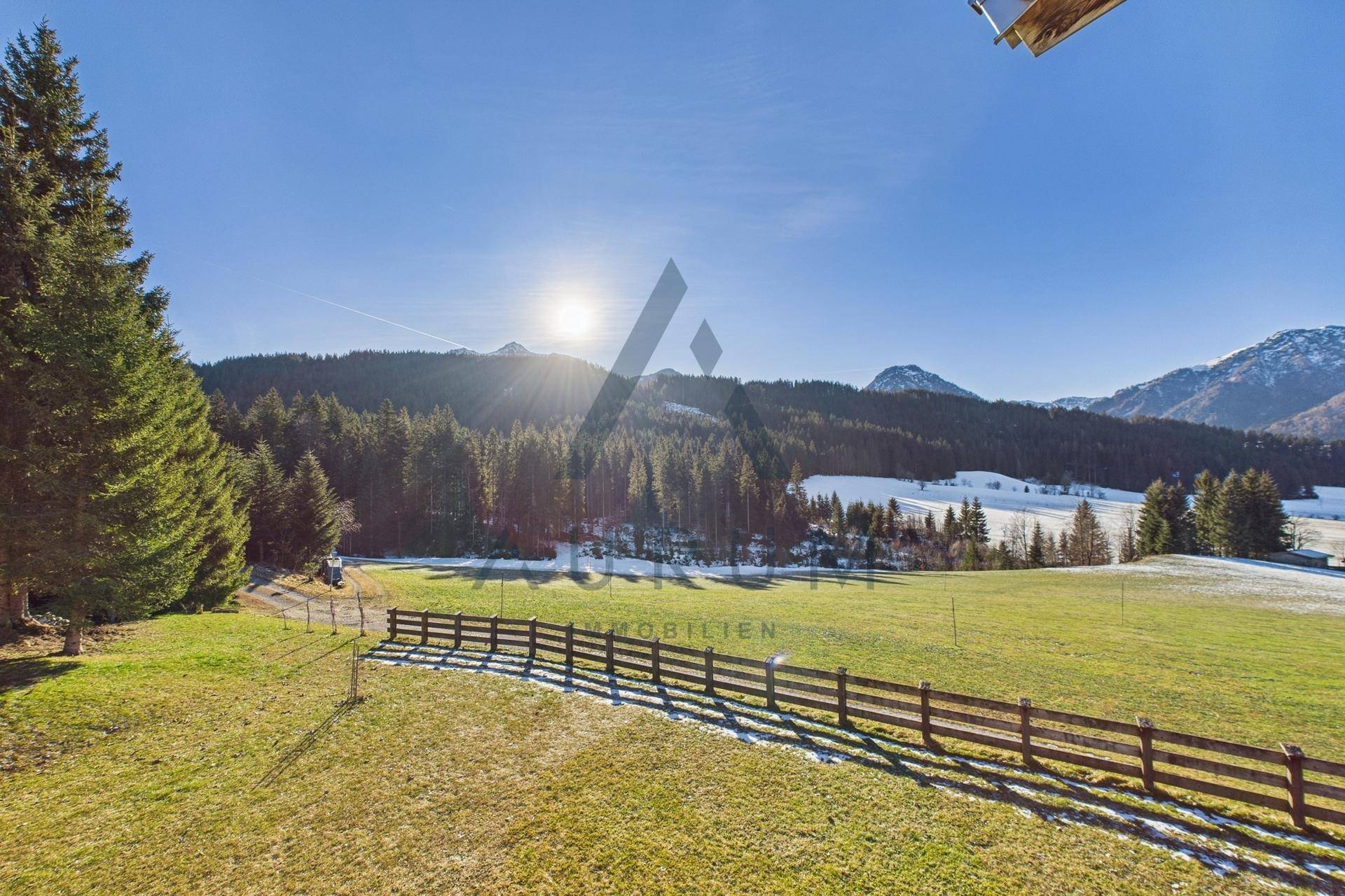 Sonniger Ausblick auf die weite Landschaft mit Wald, Wiesen und schneebedeckten Bergen.