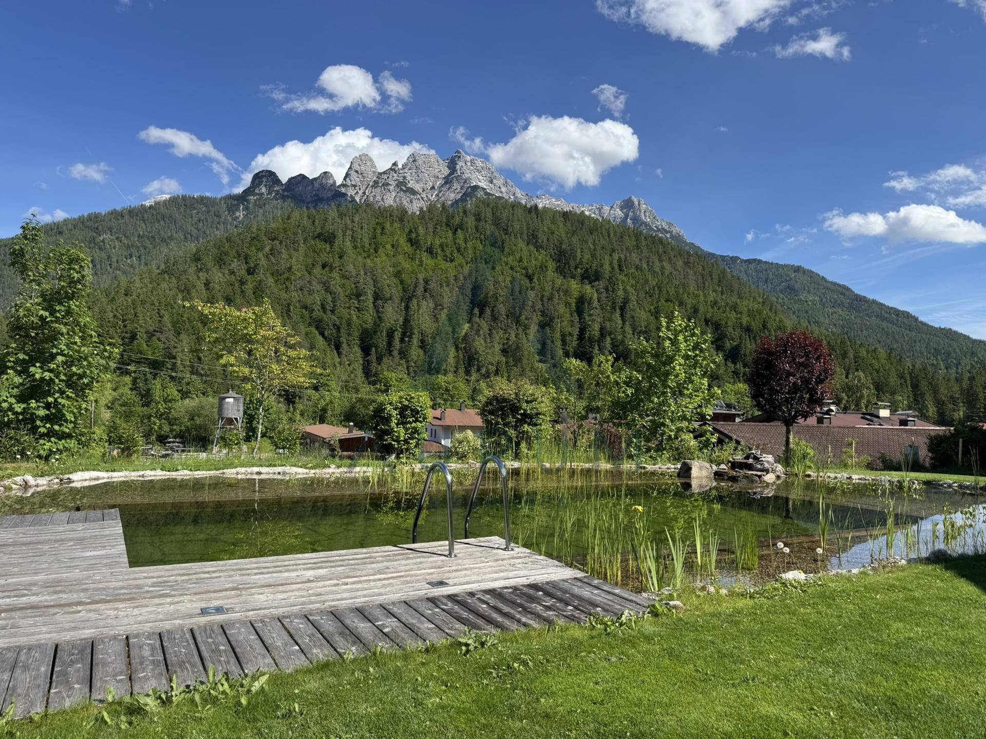 Idyllischer Garten mit Naturteich, Holzsteg und Blick auf die bewaldeten Berge.