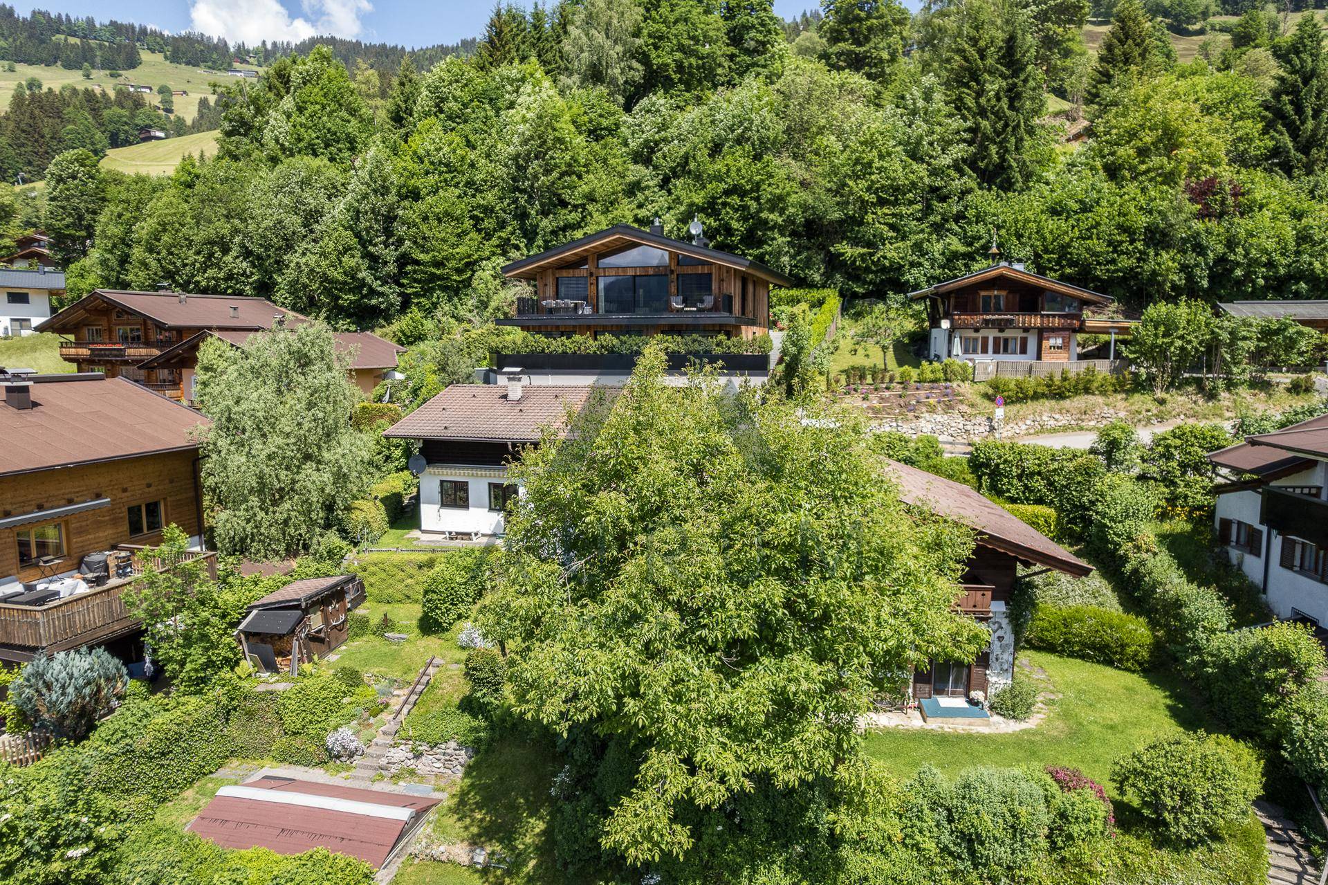 Luftaufnahme eines Wohngebiets mit Häusern, Gärten und bewaldeten Hügeln im Hintergrund.