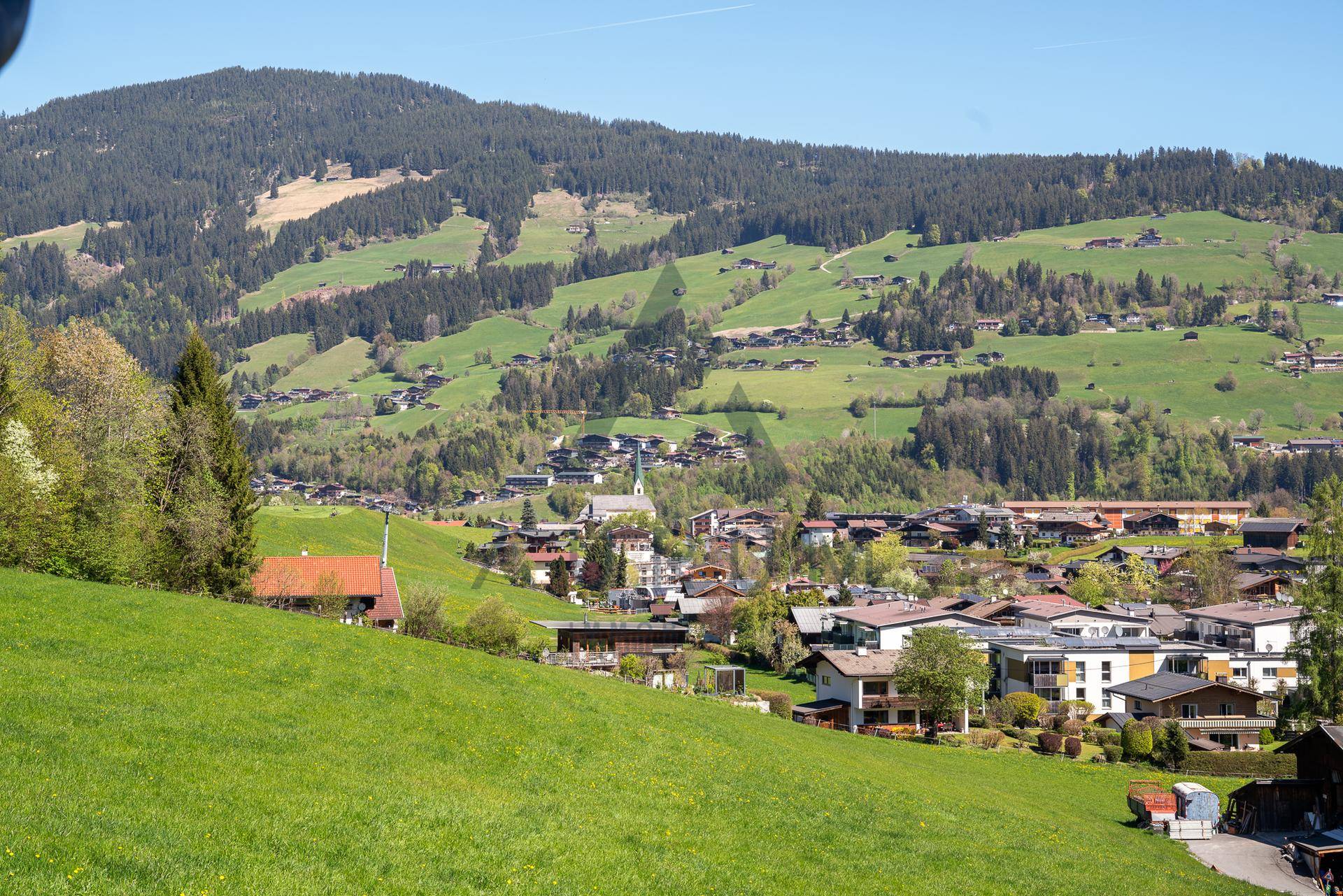 Weitläufiger Blick über ein idyllisches Bergdorf mit grünen Hügeln und bewaldeten Bergen im Hintergrund.