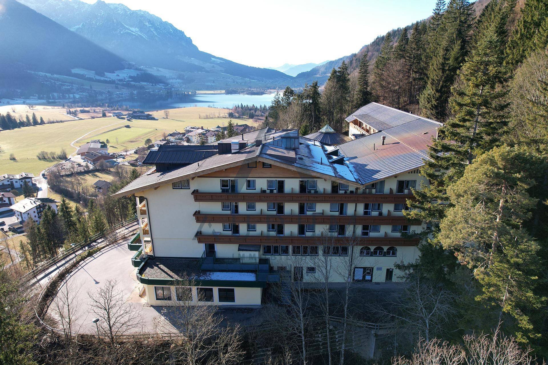 Luftaufnahme des Gebäudes mit Blick auf die Dächer, Balkone und die umliegende Berglandschaft mit einem See im Hintergrund.