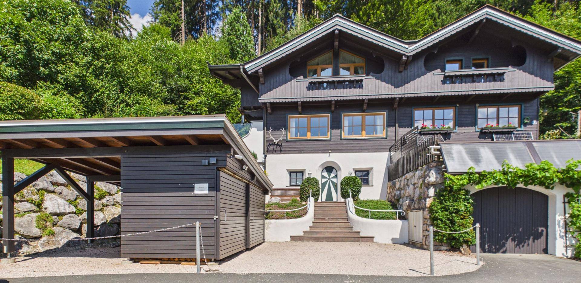Vorderansicht des Hauses mit Eingangstreppe, Carport und Garage, umgeben von viel Grün.