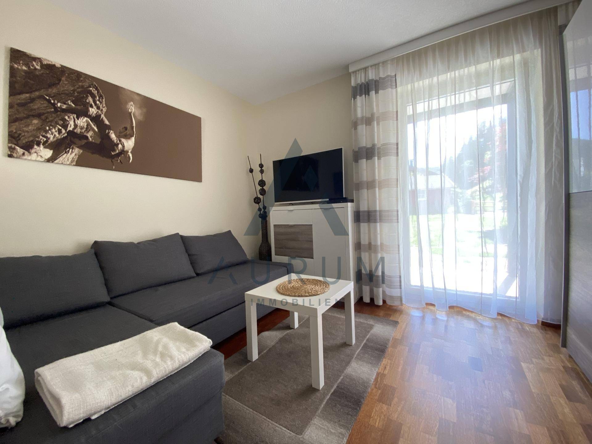 Helles Wohnzimmer mit Sofa, Fernseher und Zugang zu einem Balkon oder Garten.