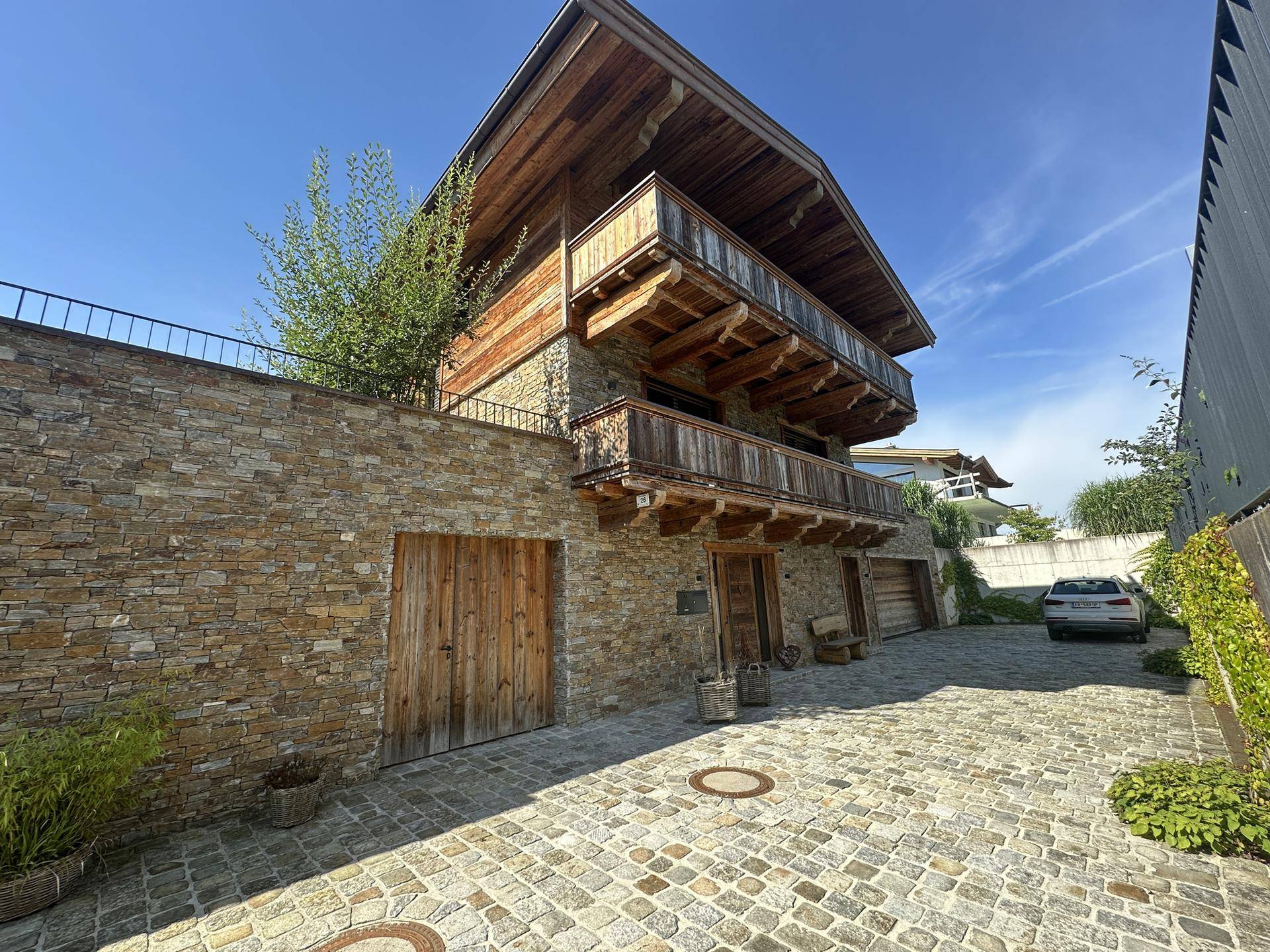 Elegante Steinfassade des Hauses mit Holzelementen und gepflasterter Auffahrt, umgeben von Natur.