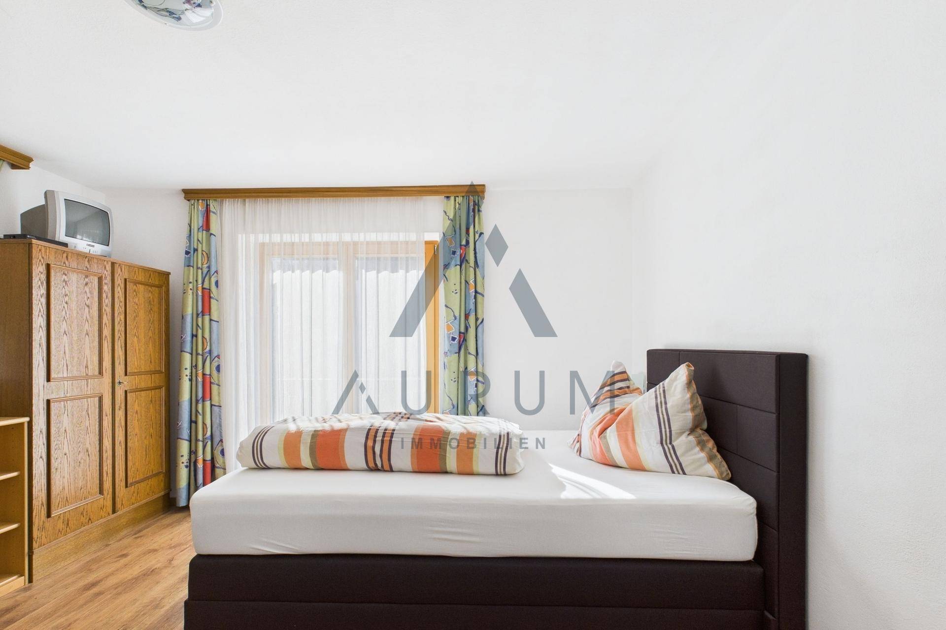 Schlafzimmer mit Doppelbett, Holzschrank und Fenster mit bunten Vorhängen.