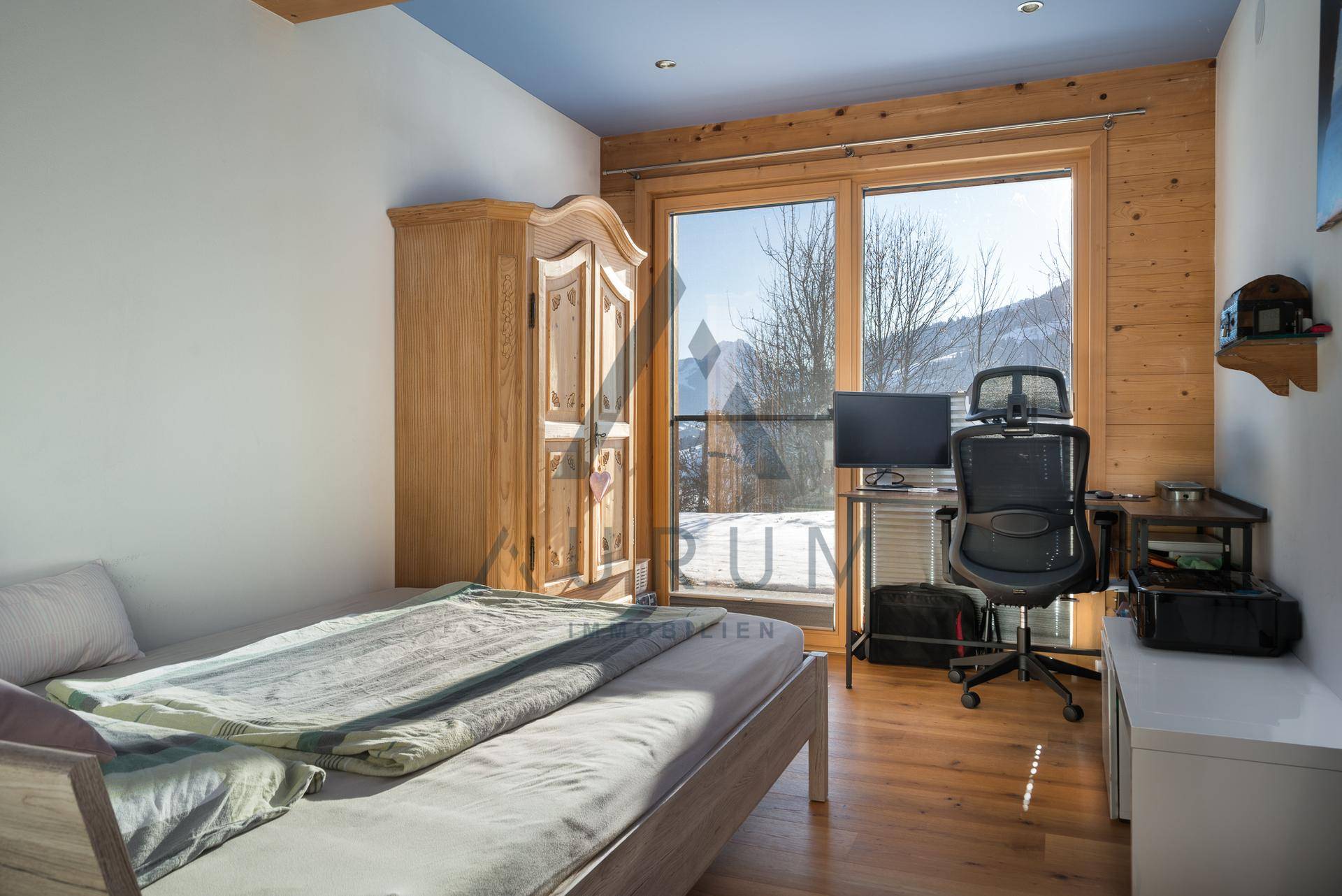 Schlafzimmer mit Holzboden, Schreibtisch und großen Fenstern mit Blick auf die verschneite Landschaft.