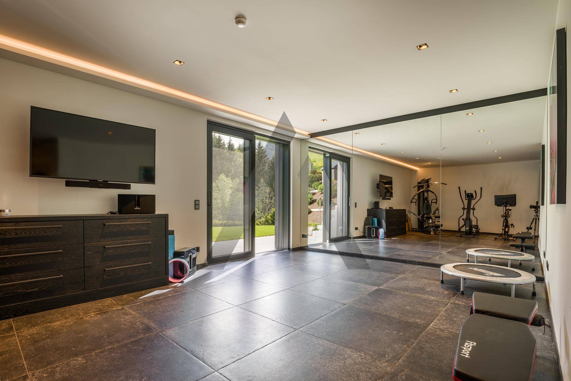 Geräumiger Fitnessraum mit Spiegelwand, Sportgeräten und Zugang zum Garten.