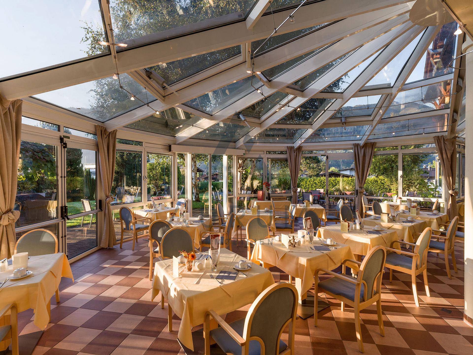 Heller Wintergarten mit vielen Tischen, kariertem Fliesenboden und Glasdach, ideal für Mahlzeiten.