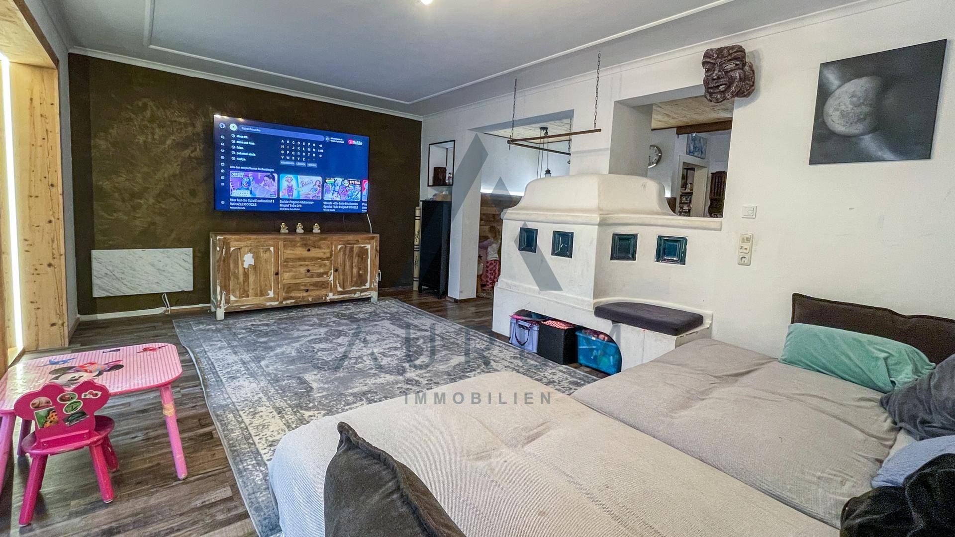 Gemütliches Wohnzimmer mit großem Teppich, Fernseher und traditionellem Kachelofen.